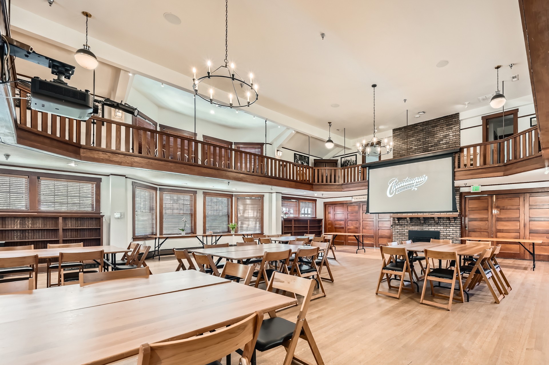 A grand two-story hall set up for events with wooden tables, folding chairs, an upper mezzanine balcony, iron ring chandeliers, and a large projection screen over a brick fireplace.