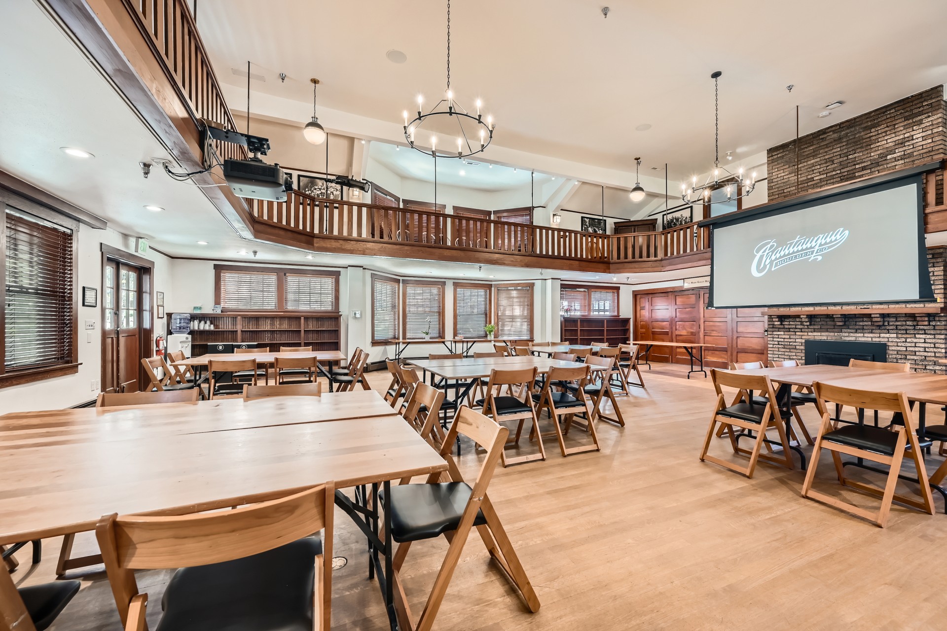 An alternate perspective of the wooden event hall, highlighting light wood tables with black-cushioned chairs, a wraparound balcony mezzanine, and a brick fireplace presentation area.