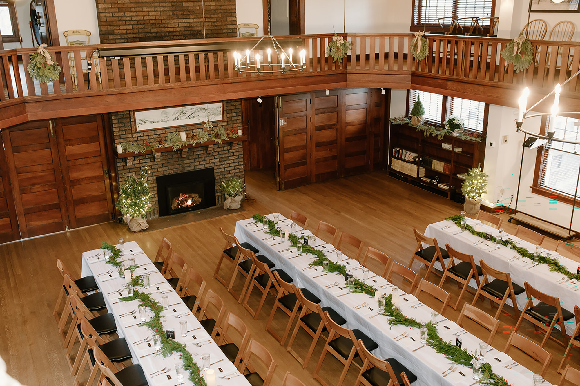 An elevated view of a spacious lodge-style banquet hall with long banquet tables set for a event, detailed with evergreen garlands, framed by wooden wall paneling, a brick fireplace, and an overhead wooden mezzanine railing.
