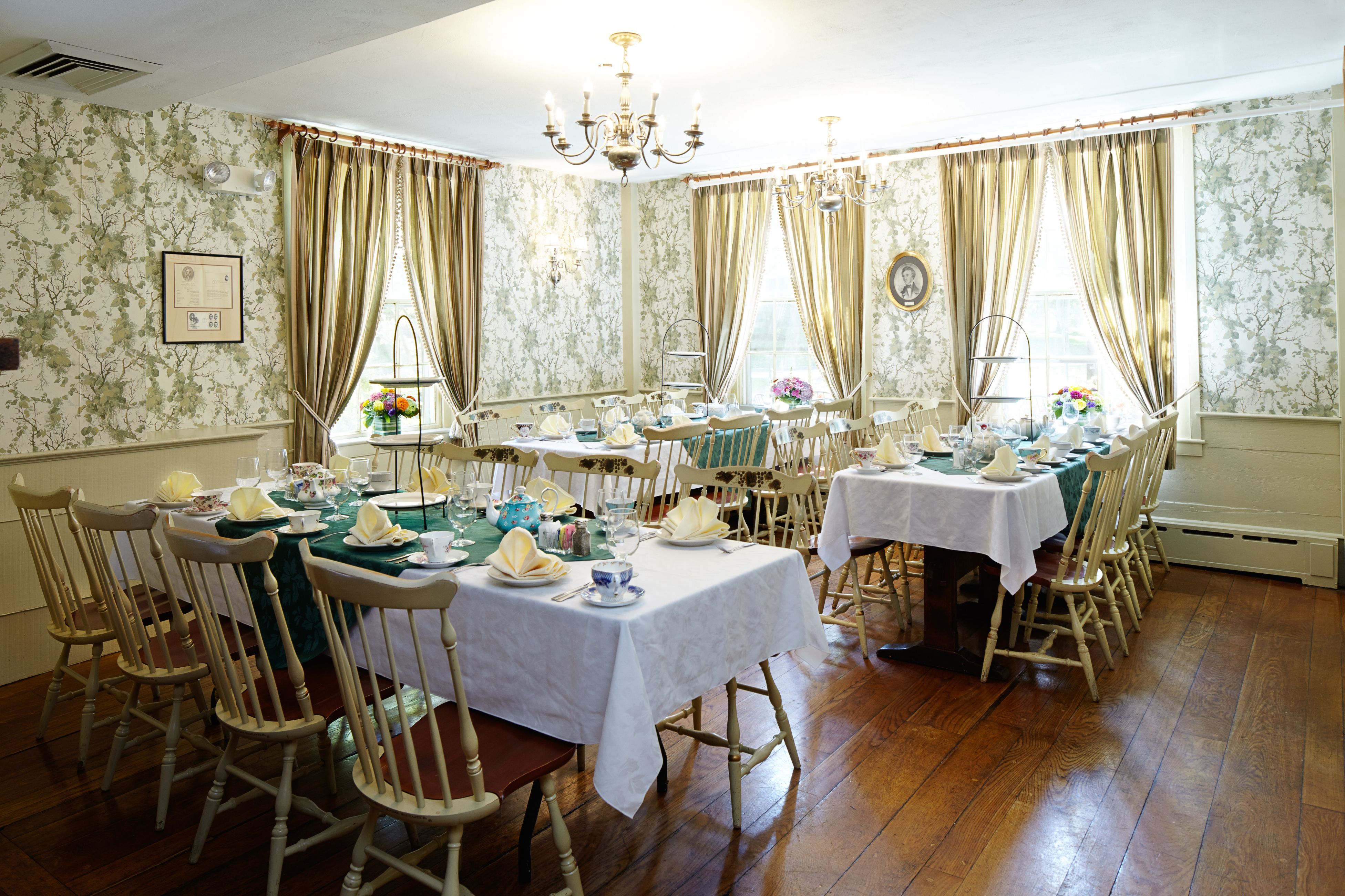 A tea room featuring light floral wallpaper, wooden floors, brass chandeliers, and rectangular tables set with white tablecloths, green table runners, teacups, and tiered serving stands.