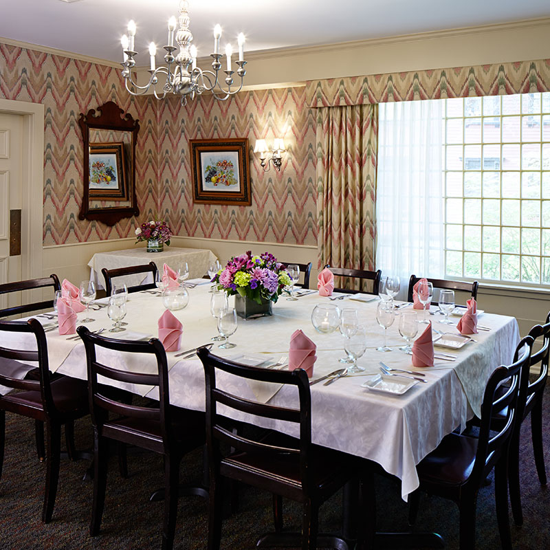 A private dining room set with a large square table, white tablecloth, folded pink napkins, a floral centerpiece, patterned wallpaper, and a silver chandelier.