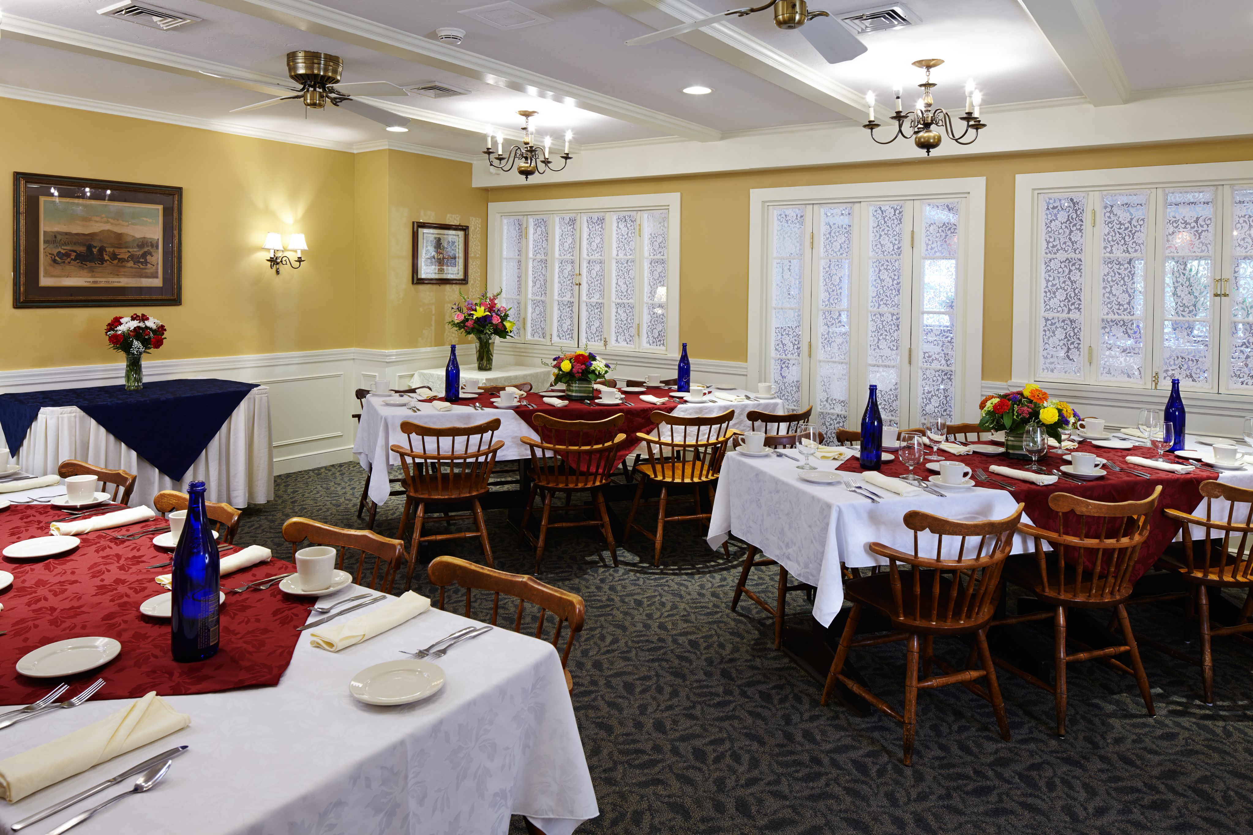 A warm-toned event room with yellow walls, white wainscoting, carpeted floor, and rectangular tables set with red and white tablecloths, wooden chairs, blue glass bottles, and floral centerpieces.