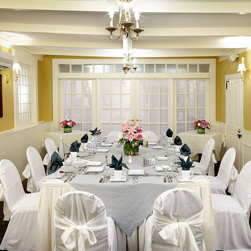 A banquet setup in a room with yellow walls, white trim, and chandeliers, featuring a large central table draped in light blue cloth, white chair covers tied with bows, and dark green fan-folded napkins.