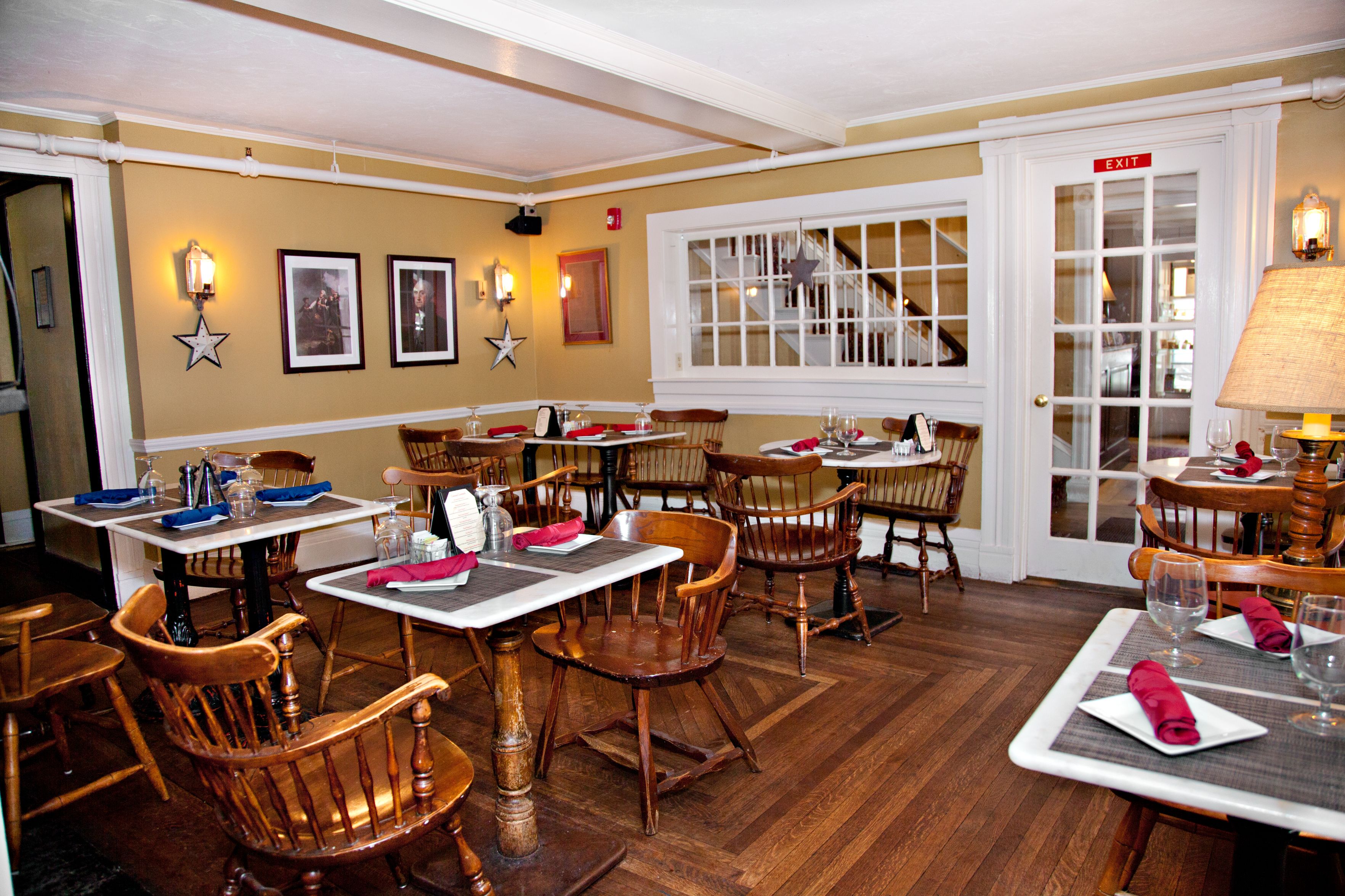 A cozy, historic-style dining room with warm beige walls, wooden Windsor chairs, small white tables with placemats and folded napkins, and framed portraits on the wall.