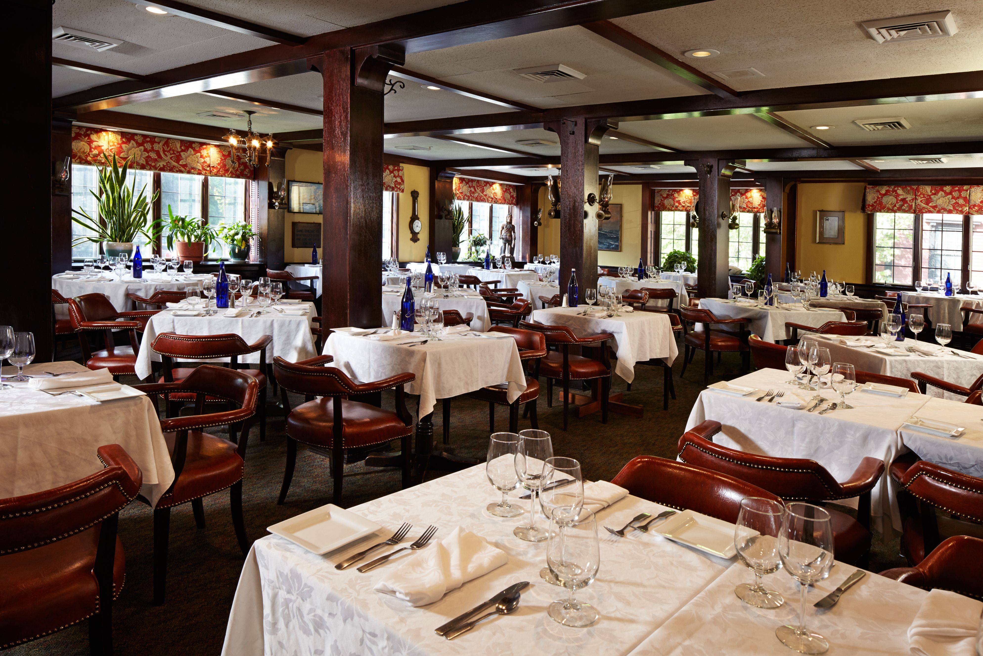 A traditional dining room featuring dark wood ceiling beams and support columns, white-clothed tables with wine glasses and settings, reddish-brown leather armchairs, and red patterned window valances.