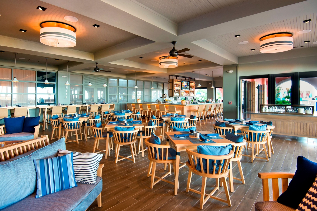 Spacious coastal dining area featuring light wood tables, chairs with bright blue cushions, casual lounge seating, and a full-service bar in the background.