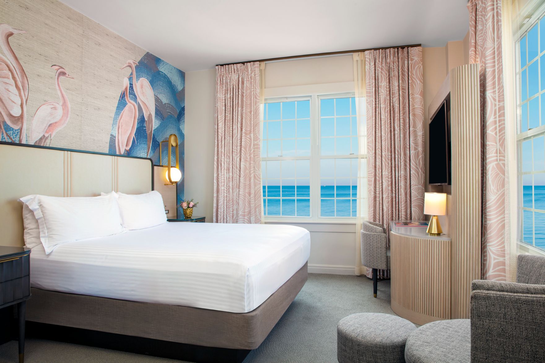 Bright, stylish hotel room featuring a king bed, pink flamingo wallpaper accent wall, blush patterned curtains, and windows overlooking the ocean.