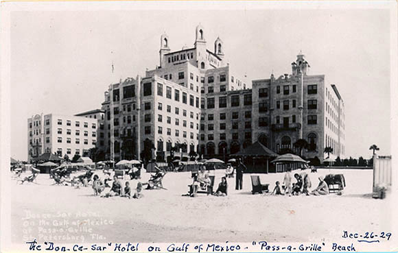 Vintage black-and-white postcard showing the historic Don CeSar Hotel on Pass-a-Grille Beach with guests relaxing on the sand in front.
