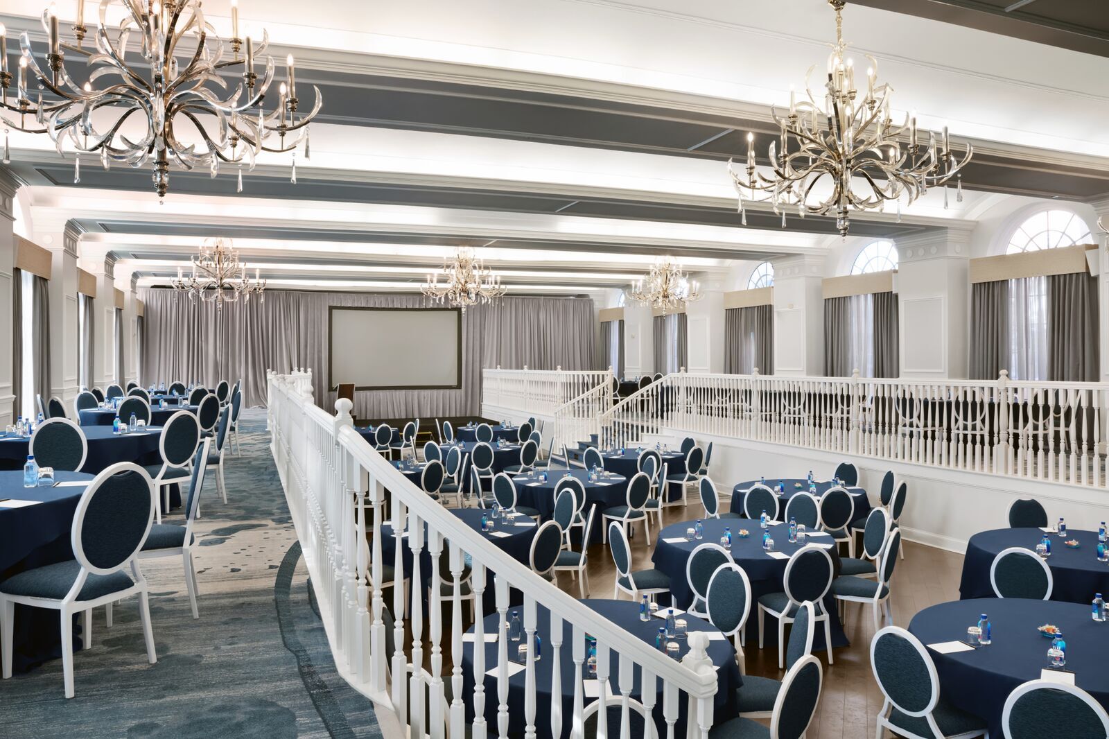 Large multi-level conference ballroom set up with round tables draped in navy blue tablecloths, white oval-backed chairs, and ornate chandeliers overhead.