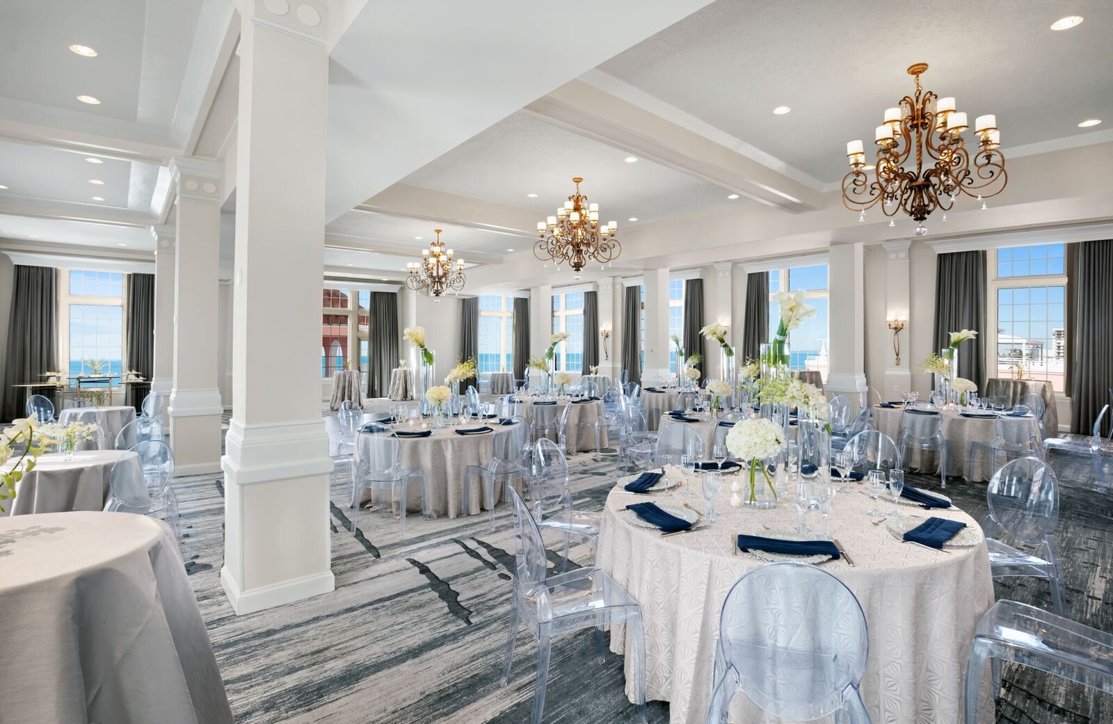 Elegant ballroom decorated for an event, featuring round tables with white linens, clear acrylic chairs, tall floral centerpieces, and chandeliers overhead.