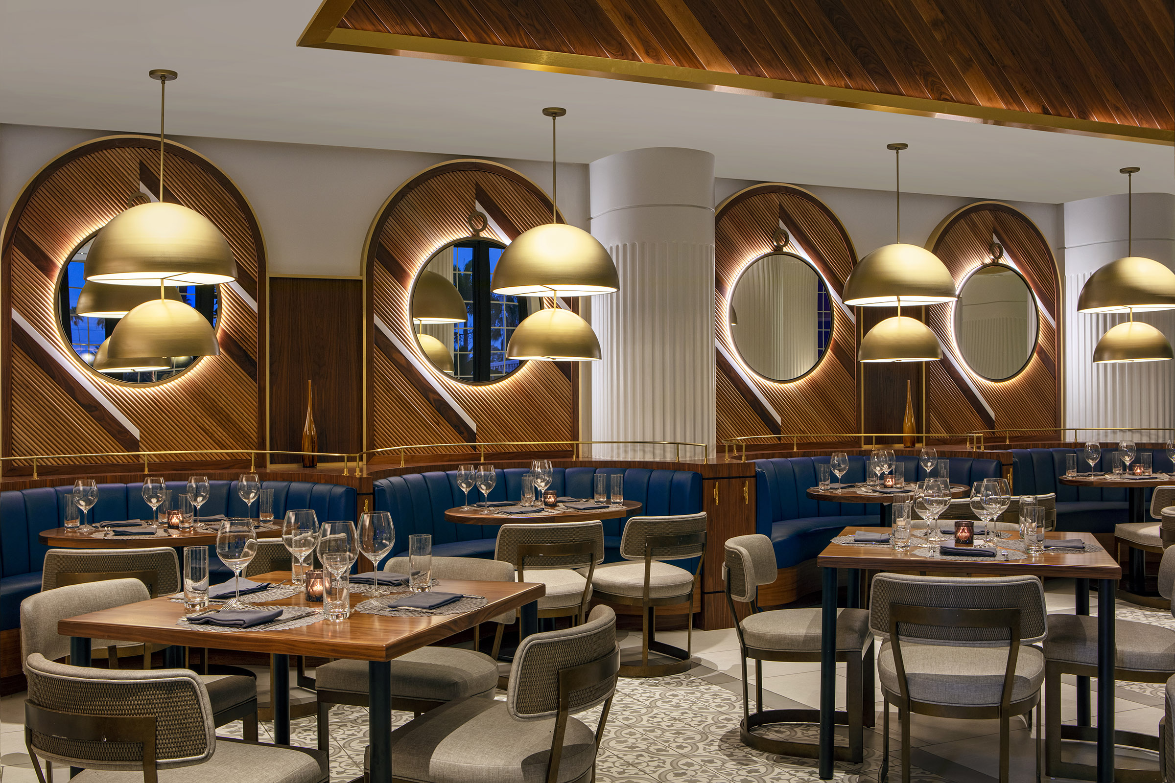 Upscale restaurant dining area featuring blue leather booth seating, warm wood-paneled walls with backlit circular mirrors, dome-shaped gold pendant lights, and neatly set tables with wine glasses.