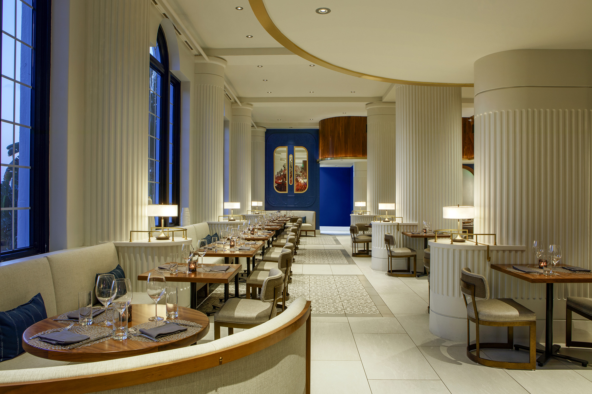 Modern upscale hotel restaurant with fluted white columns, neutral banquette seating, wooden dining tables set with wine glasses, and blue accent walls.