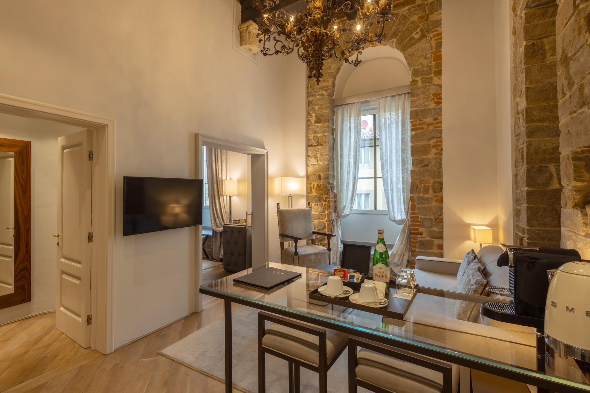Spacious suite sitting area with a glass-topped table, sofa, ornate chandelier, wall-mounted TV, and high stone archway surrounding a curtained window.