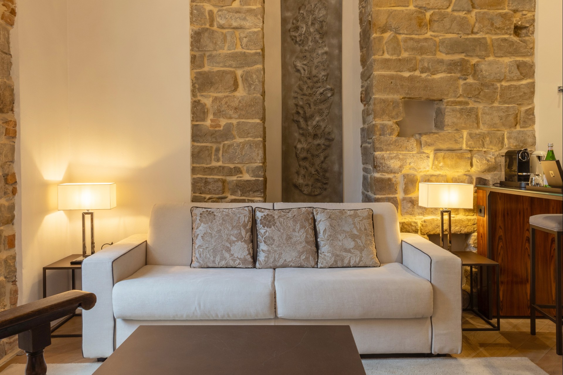 Cozy hotel living area with a beige three-seater sofa, pattern throw pillows, flanking table lamps, and exposed stone columns.