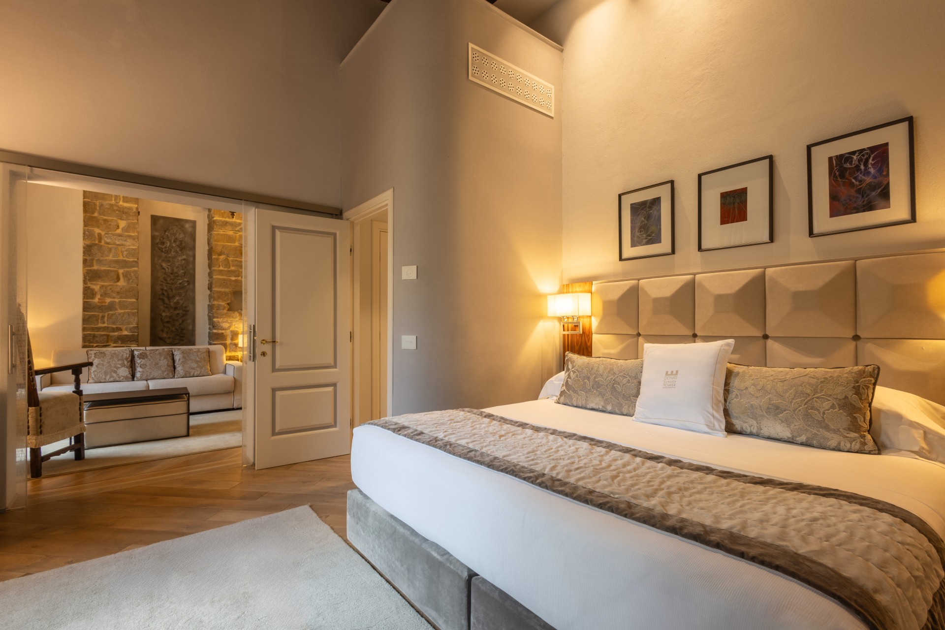 Hotel bedroom suite featuring a king-size bed with light-colored padded headboard, warm lighting, and an open doorway leading into an adjoining living room.