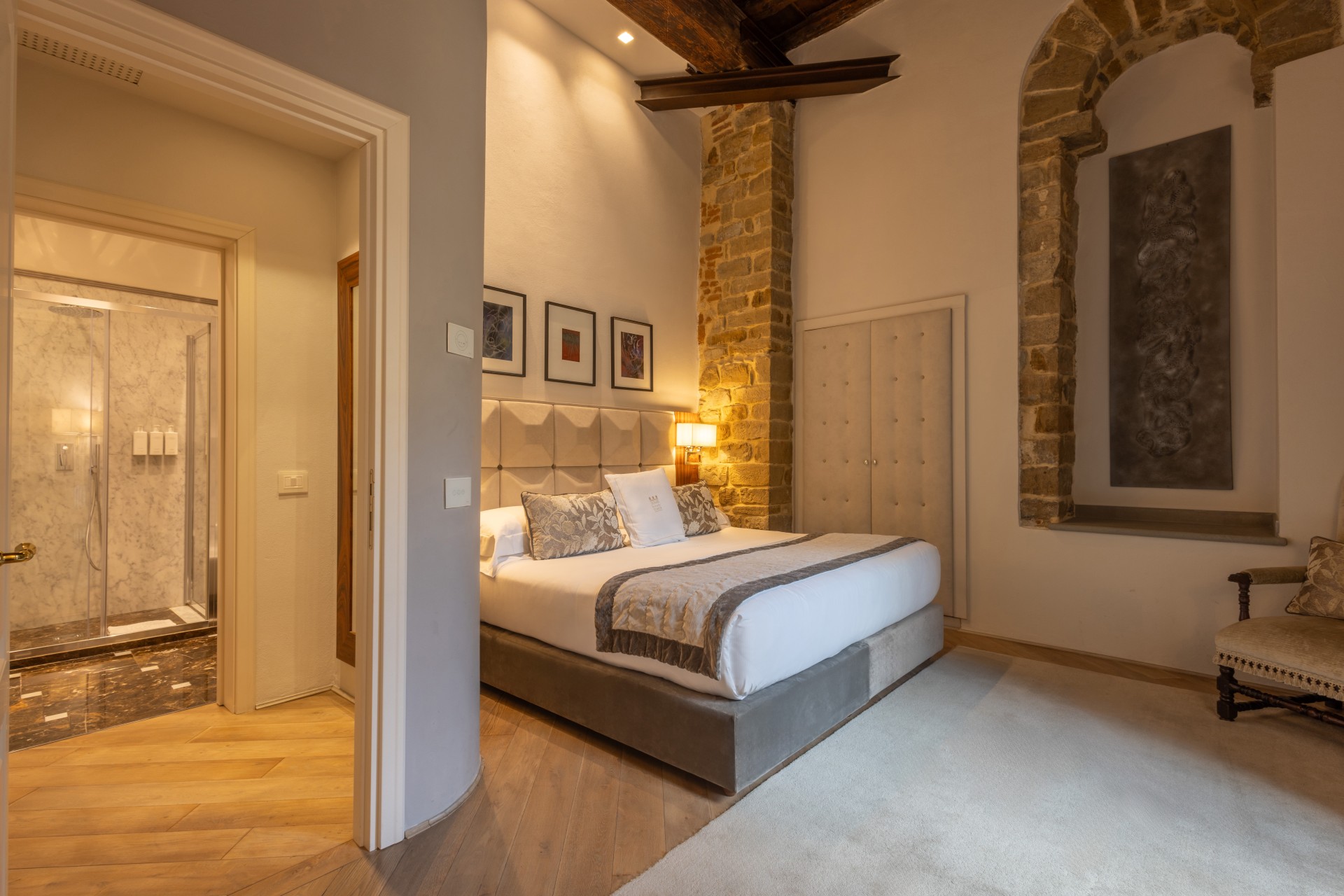 Spacious hotel bedroom with a king-size bed, upholstered tufted headboard, exposed stone columns, an adjacent marble bathroom with a glass-enclosed shower, and elegant wooden accents.