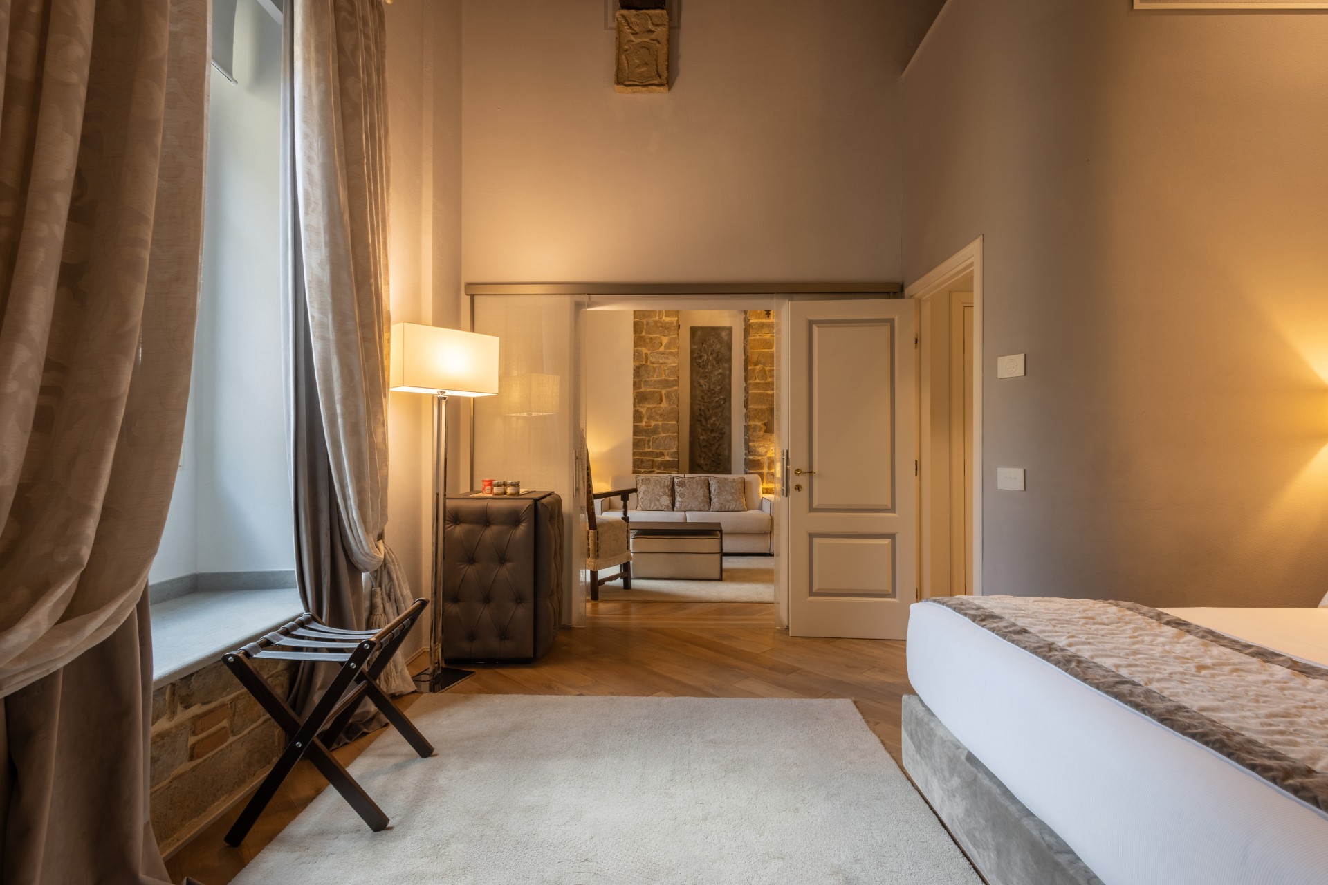 High-angle perspective of a luxury hotel bedroom featuring a bed with neutral linens, floor-to-ceiling drapes, and an open doorway revealing an adjoining living room.