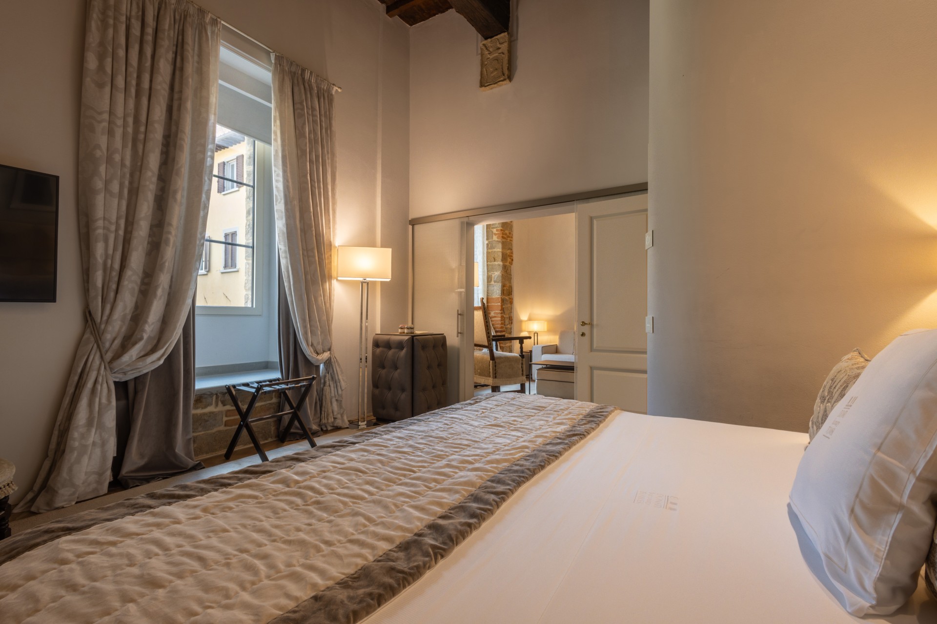 Hotel bedroom view from the bed looking toward elegant draped windows, a floor lamp, a luggage rack, and an open doorway leading to a sitting area.