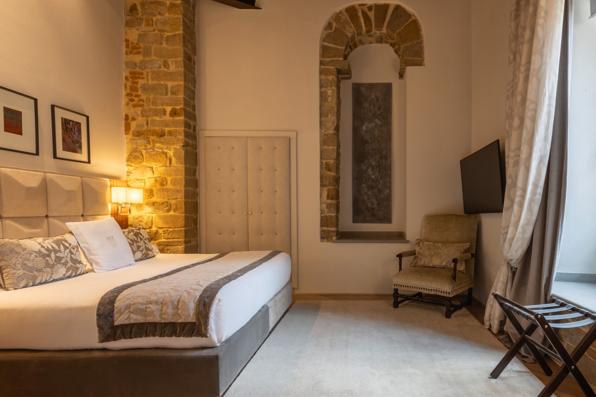 Hotel bedroom suite with a king-size bed, upholstered tufted headboard, exposed stone column and wall arch, plush armchair, and wooden luggage rack near a curtained window.