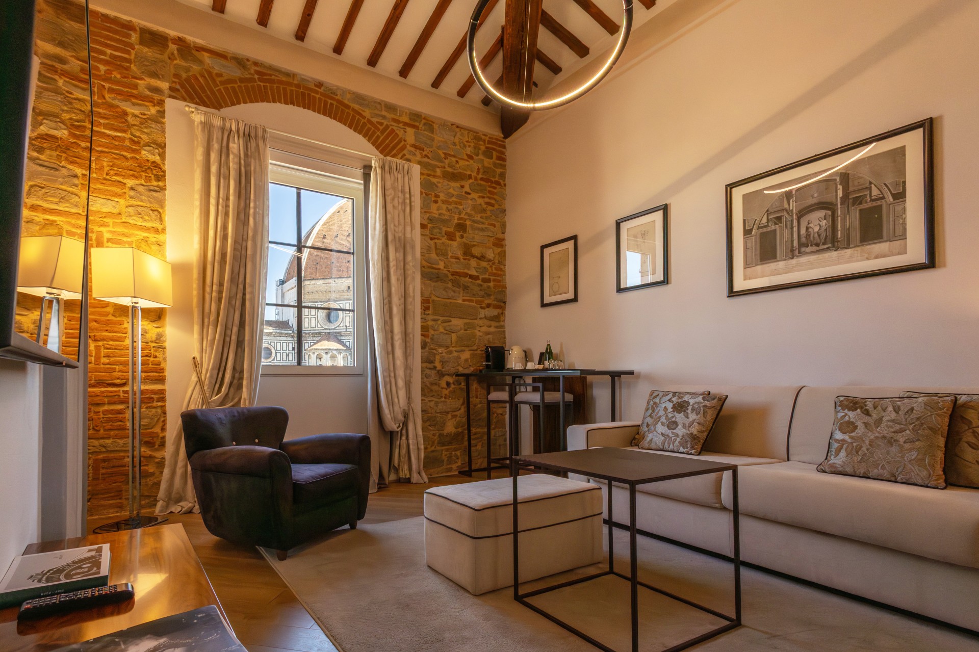 A luxury hotel suite living room with a beige sofa, exposed stone archway, circular ceiling light, and a window framed by curtains showing the Florence Cathedral dome.