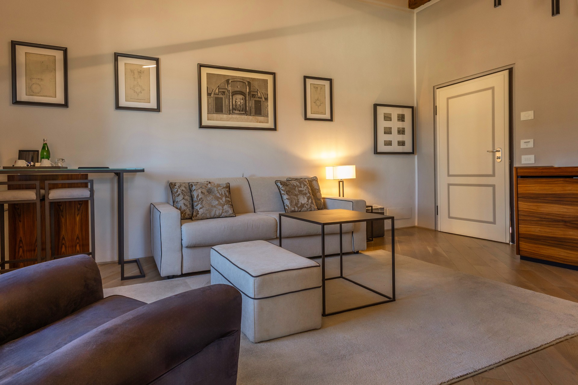 A warm, spacious hotel suite living area featuring a neutral sofa with decorative throw pillows, a matching ottoman, a modern coffee table, framed artwork on light-colored walls, and a high-top bar setup on the side.