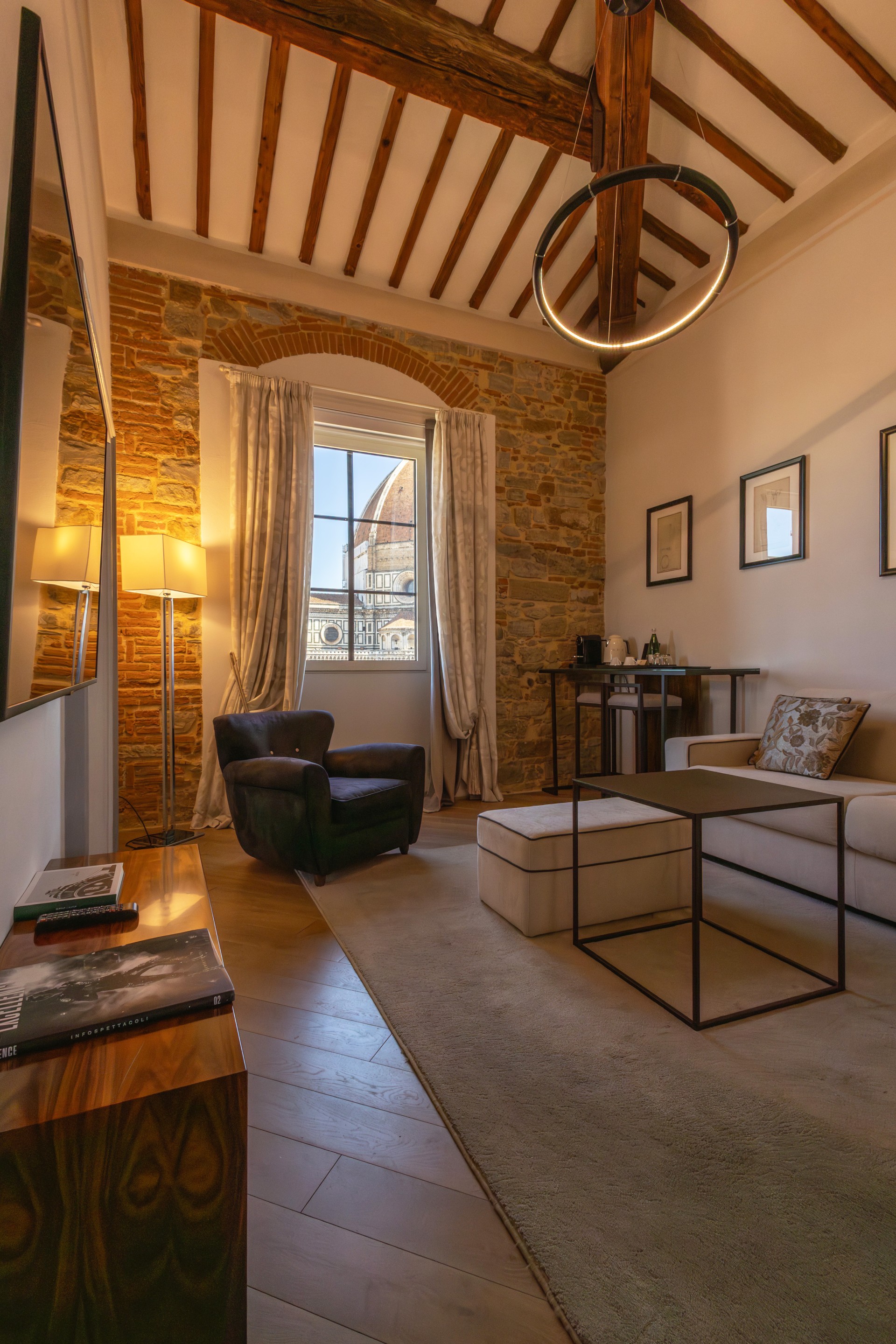 A luxury hotel suite living area featuring high vaulted wooden beam ceilings, exposed stone walls, a modern ring chandelier, and a large window offering a direct view of the Florence Cathedral dome.