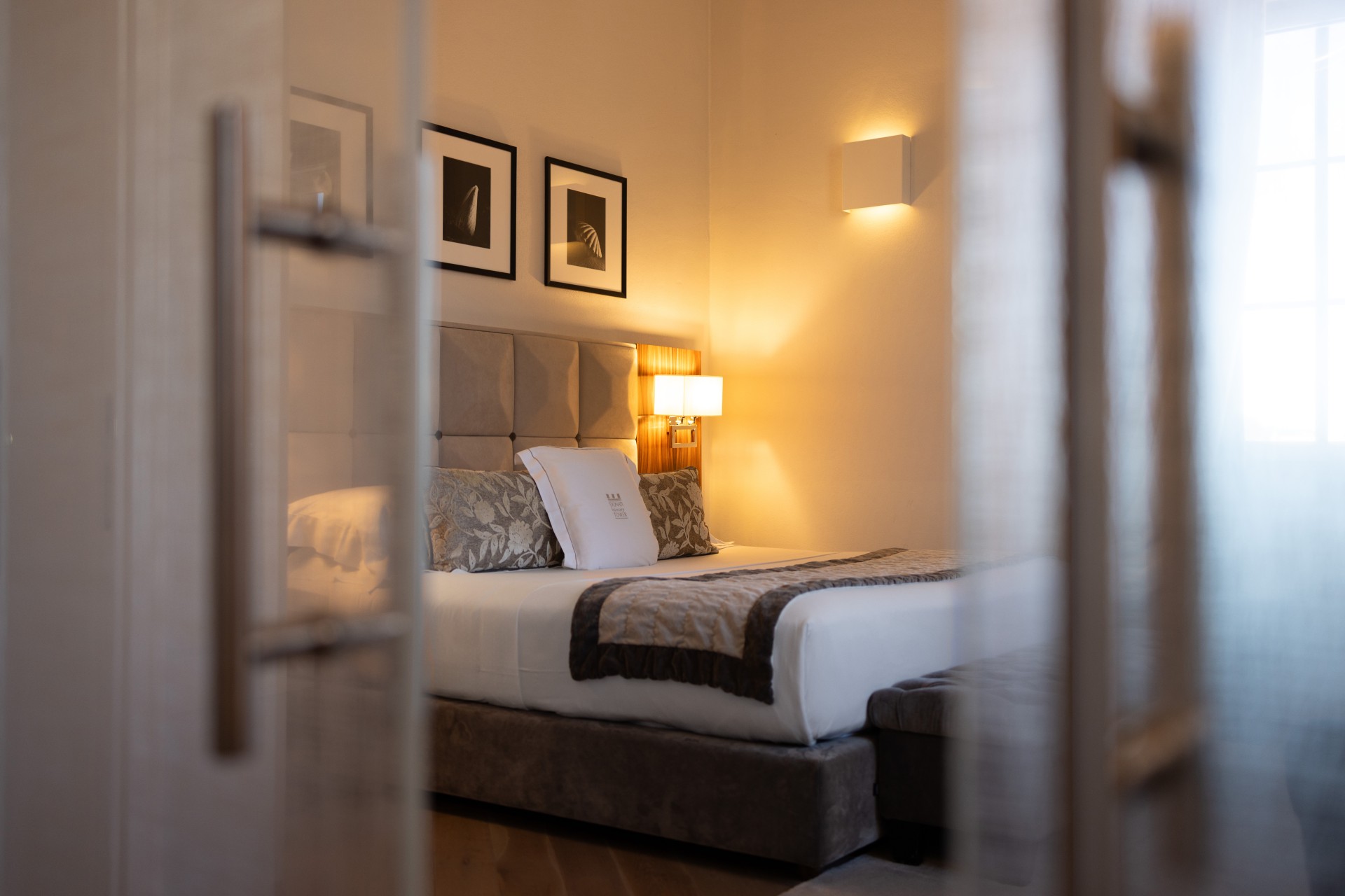 A cozy, warmly lit hotel bedroom viewed through glass double doors, featuring a king-size bed with a tufted headboard, decorative throw pillows, and framed artwork.