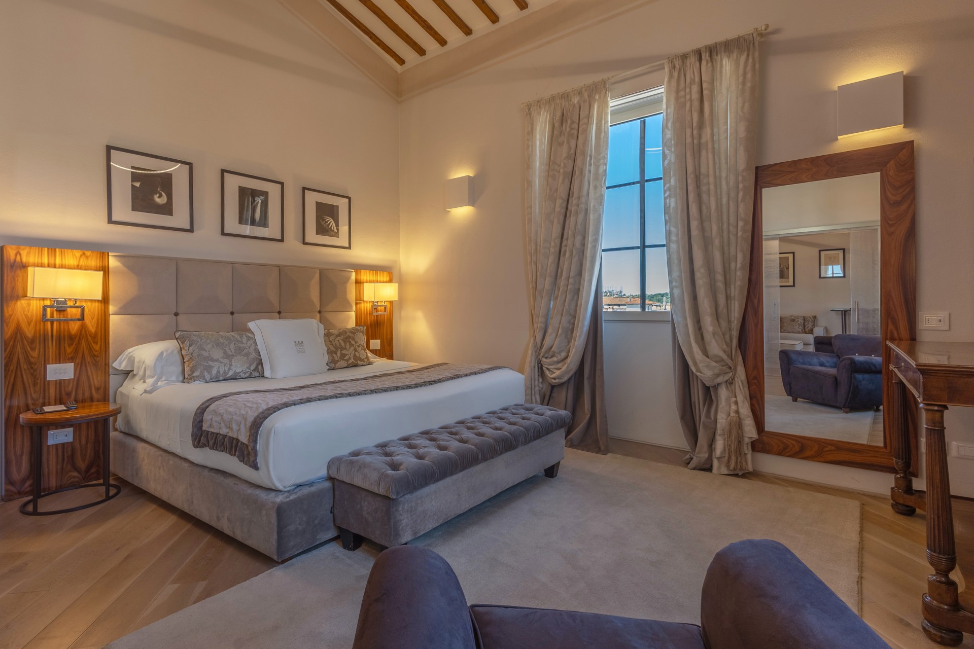 An elegant hotel bedroom featuring a plush king-size bed with a tufted headboard, a grey tufted bench at the foot of the bed, a large full-length mirror, warm lighting, and a window with light beige curtains.