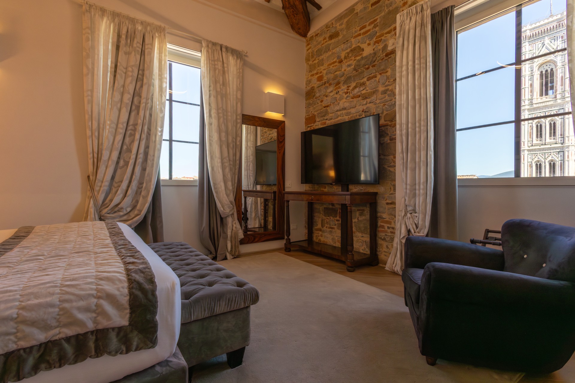 A luxury hotel bedroom featuring an exposed stone wall, a large bed with a grey tufted bench, a plush armchair, and windows providing a direct view of Giotto's Campanile in Florence.