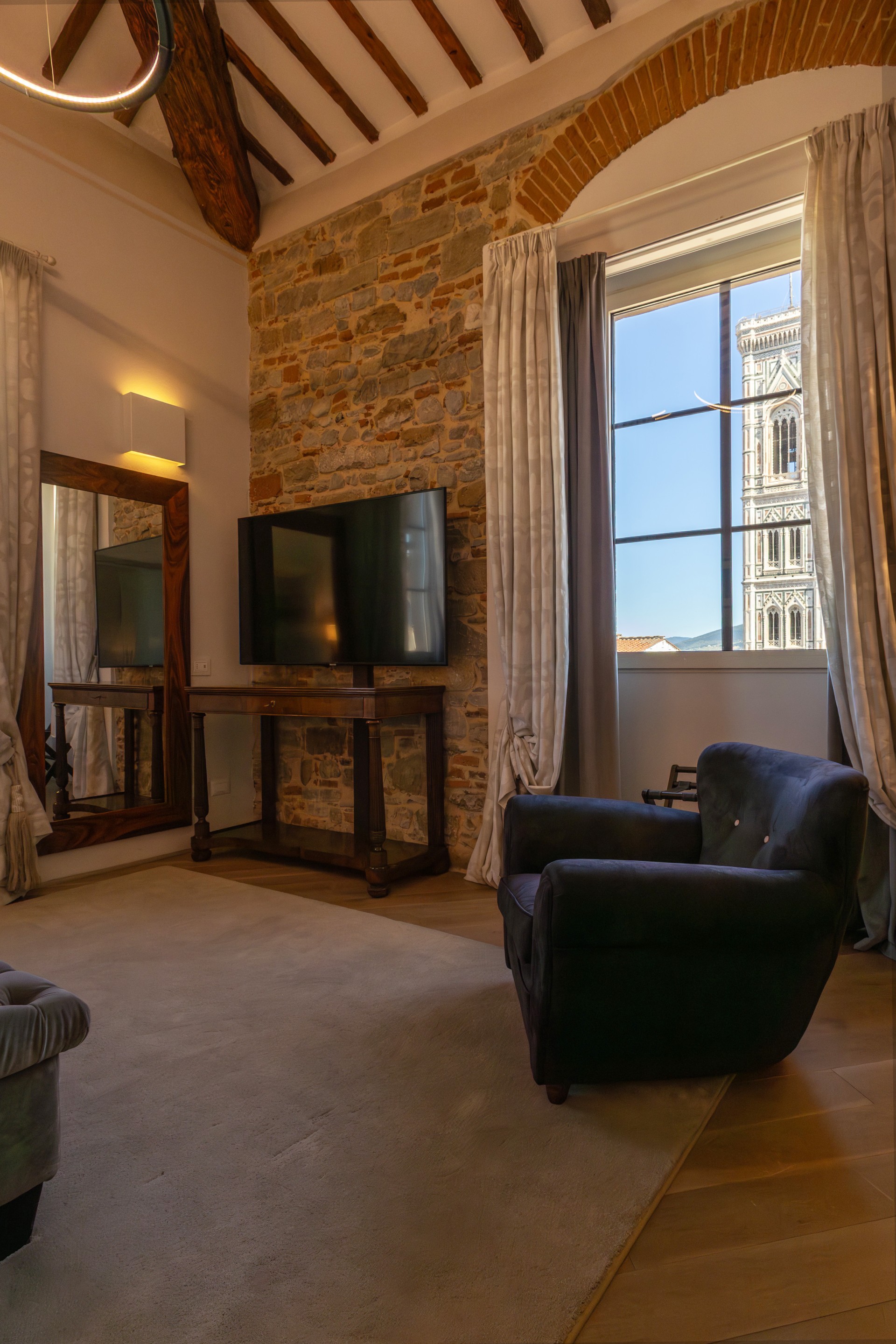 A stylish hotel suite sitting area with exposed brick walls, wooden ceiling beams, a large mirror, a comfortable armchair, and a window framing Giotto's Campanile in Florence.