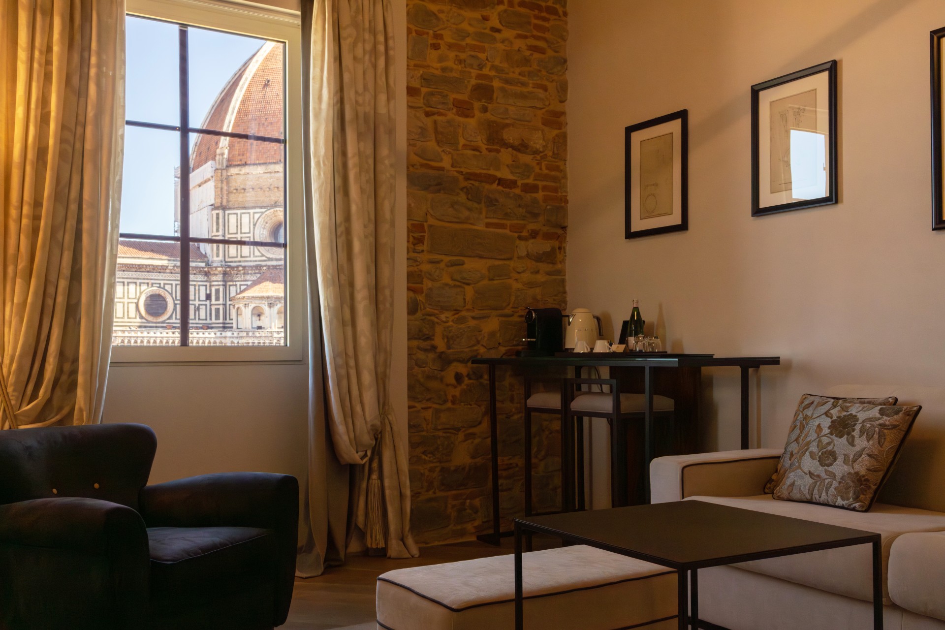 A hotel suite sitting area featuring a dark armchair, cream sofa, coffee station, and a window framed by curtains looking out at the Florence Cathedral dome.