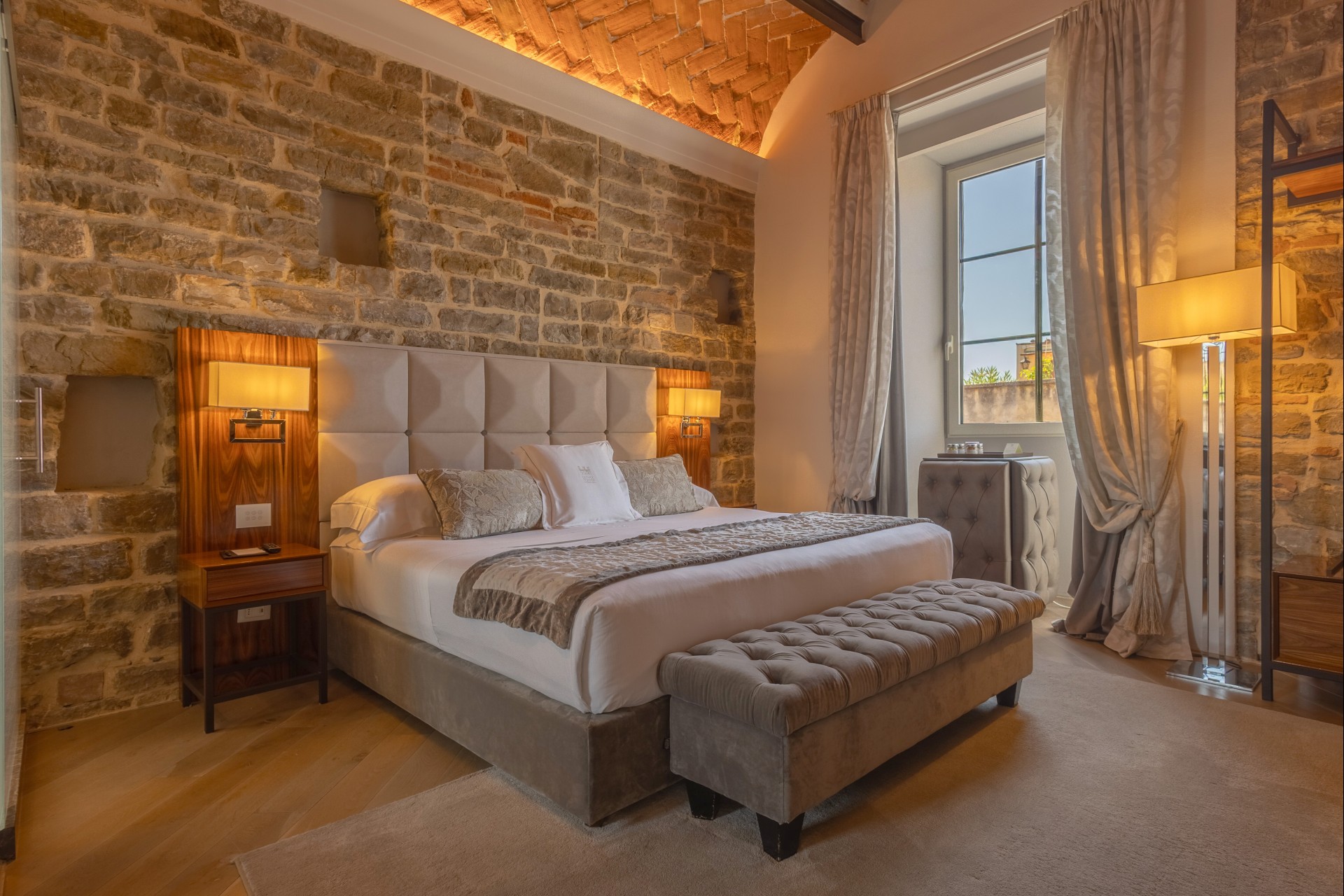 Spacious hotel bedroom with an illuminated brick vaulted ceiling, exposed stone walls, a king-sized bed with an upholstered bench at the foot, and a large window.