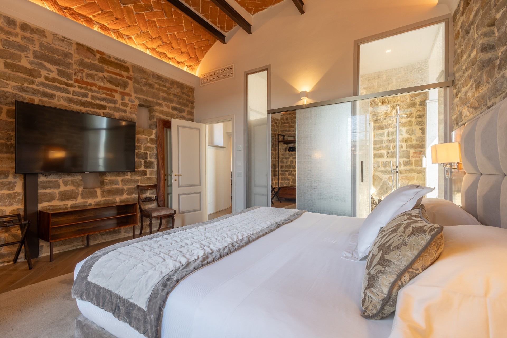 Hotel bedroom suite with warm backlit terracotta brick ceiling, exposed stone walls, large flat-screen TV mounted on the wall, and frosted glass sliding door leading to an en-suite shower area.