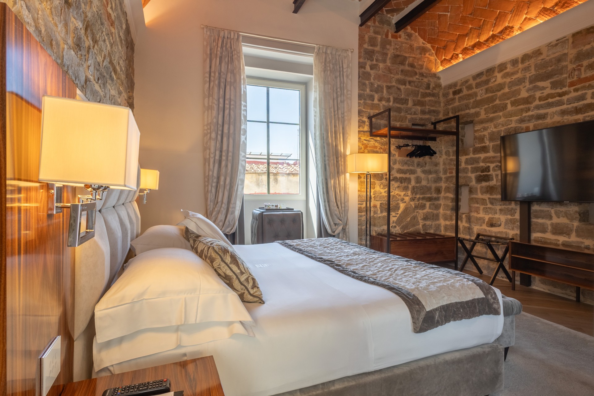 Spacious luxury hotel bedroom featuring a plush king bed, exposed stone and brick accent walls, a wall-mounted TV, and a bright window with full drapes.