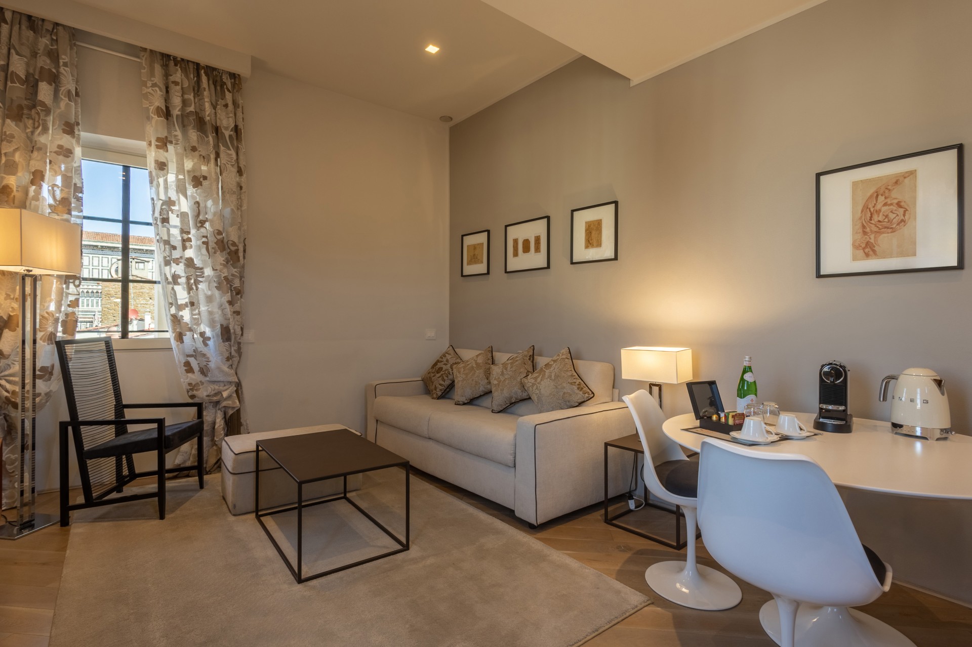 Brightly lit hotel living area with a neutral sofa, coffee table, white dining table with tulip chairs, and patterned drapes framing a window.
