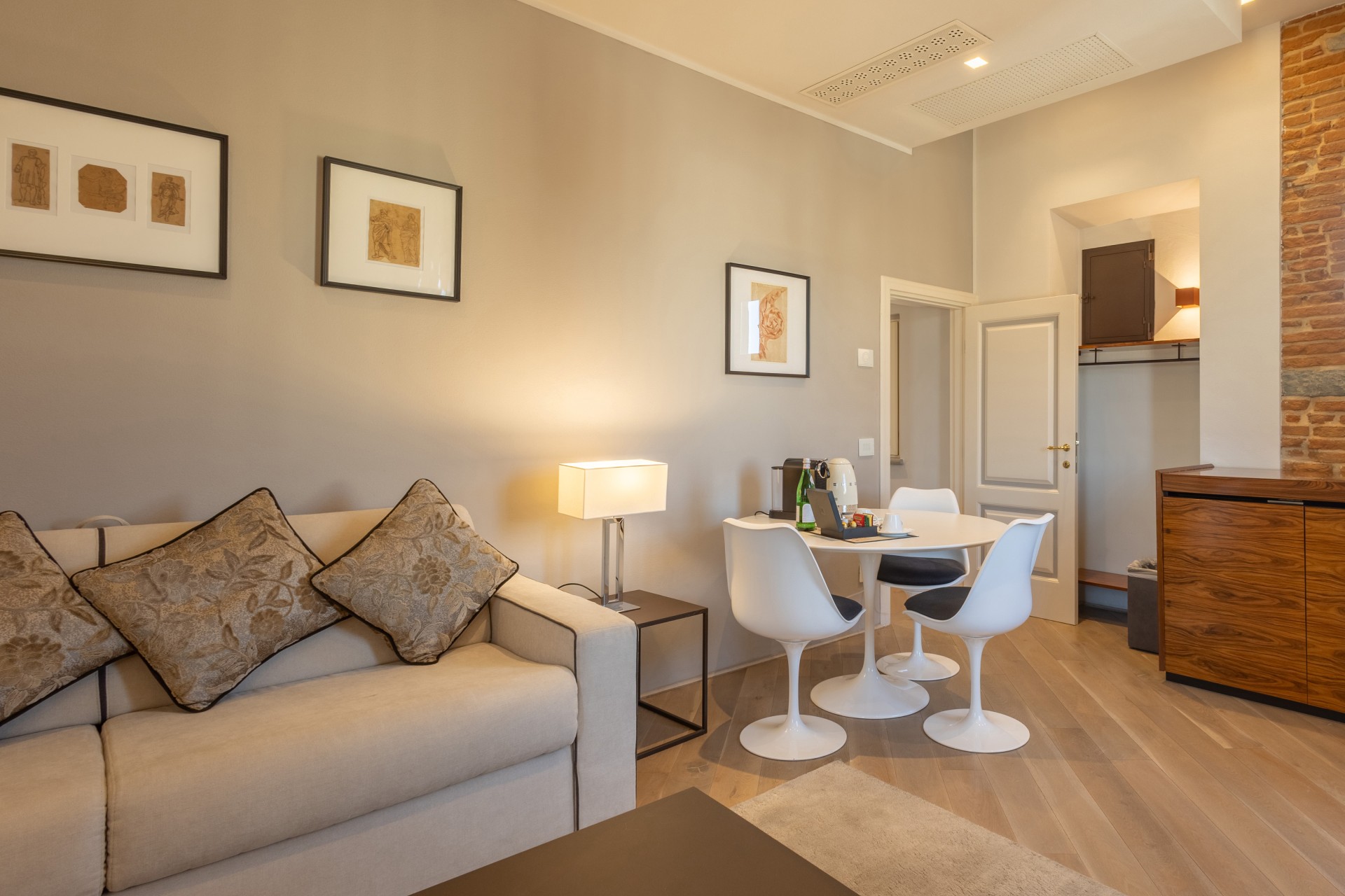 Modern hotel suite living space with a neutral-toned sofa, small dining set with tulip chairs, framed artwork on grey walls, and light hardwood floors.