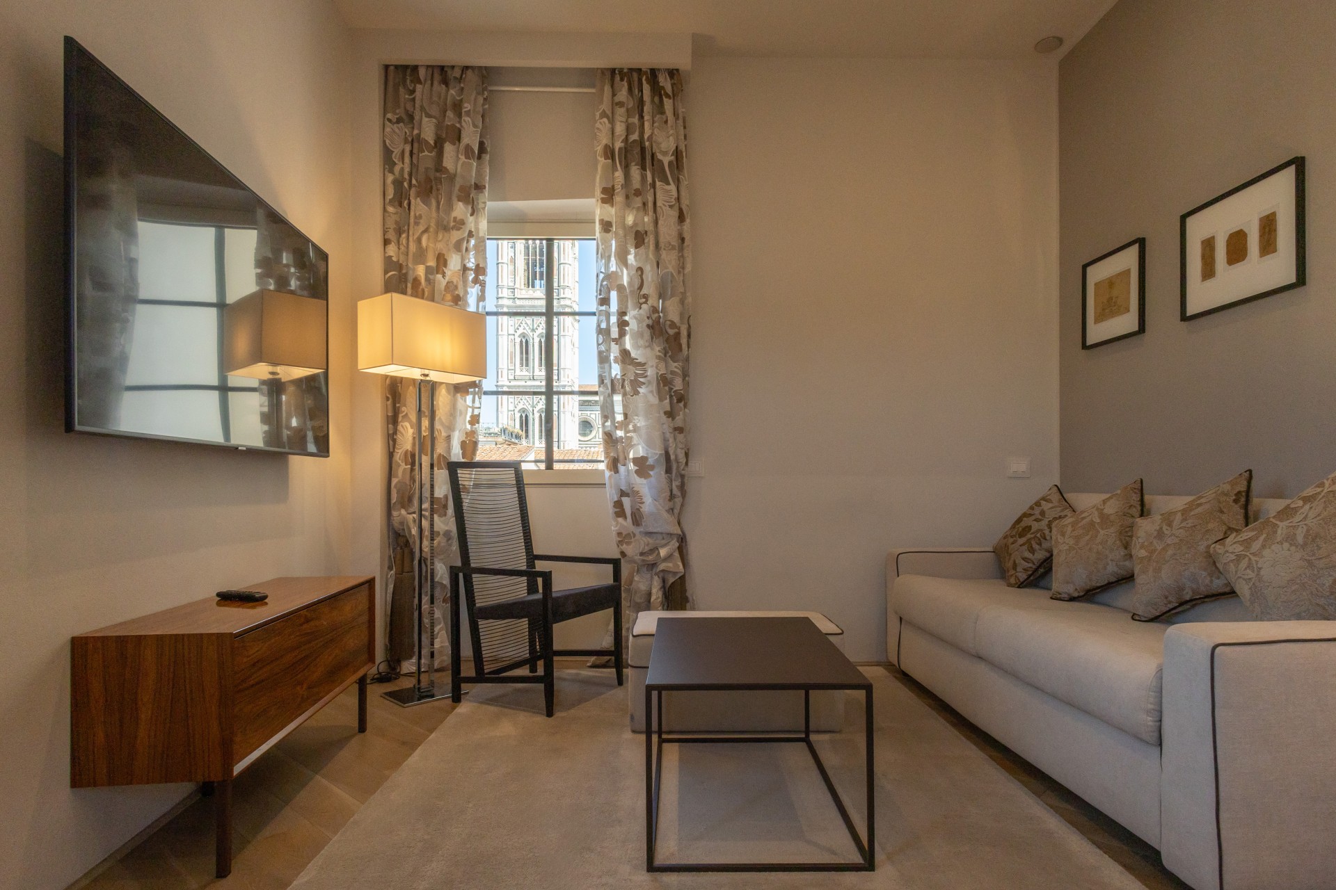 Cozy hotel sitting area with a light grey sofa, coffee table, wall-mounted TV over a wooden console, and a window framed by curtains offering a view of Giotto's Bell Tower.