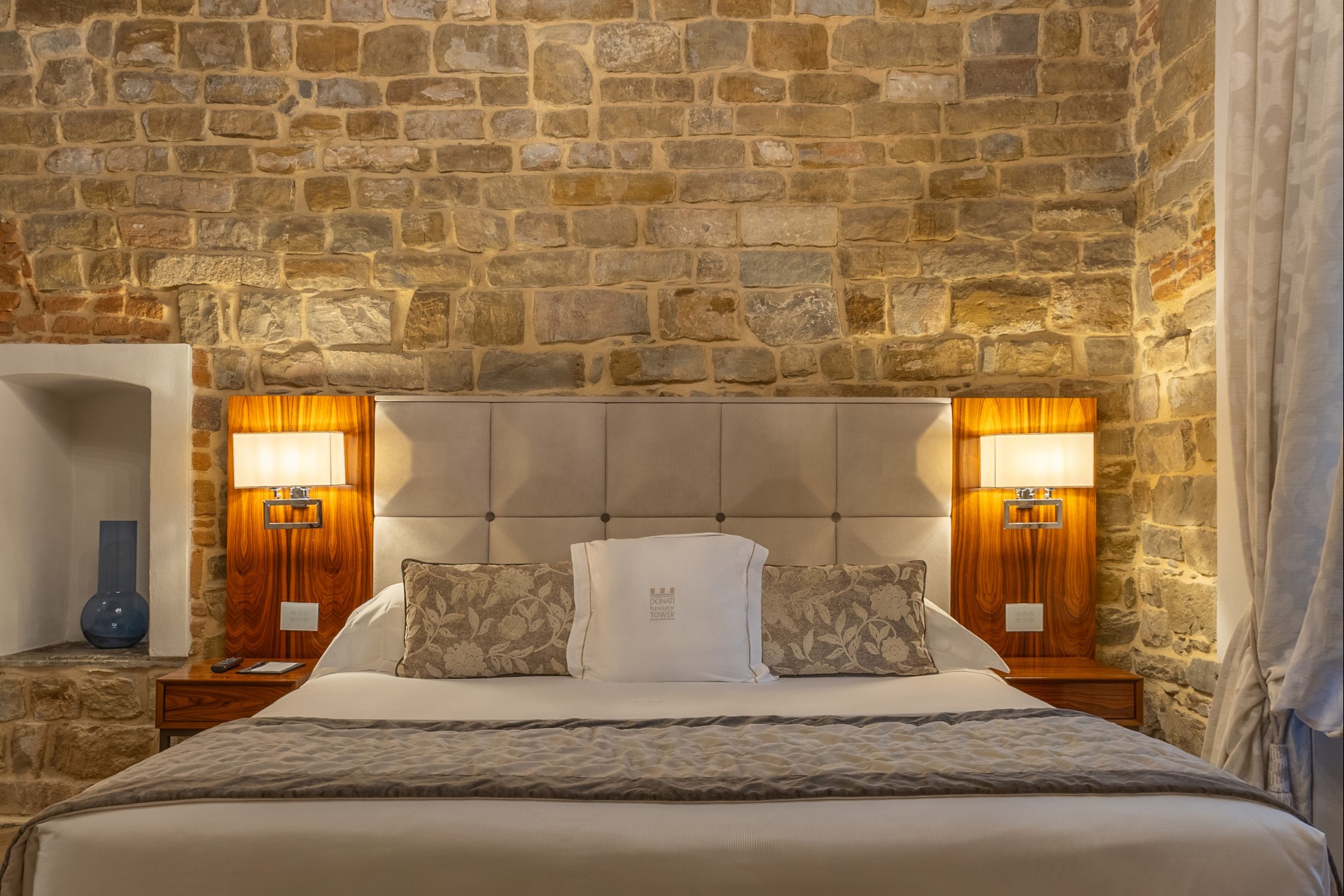 Close-up of a hotel suite bed with a tufted headboard, wood-paneled nightstands, sconce lamps, decorative pillows, and exposed stone wall backdrop.