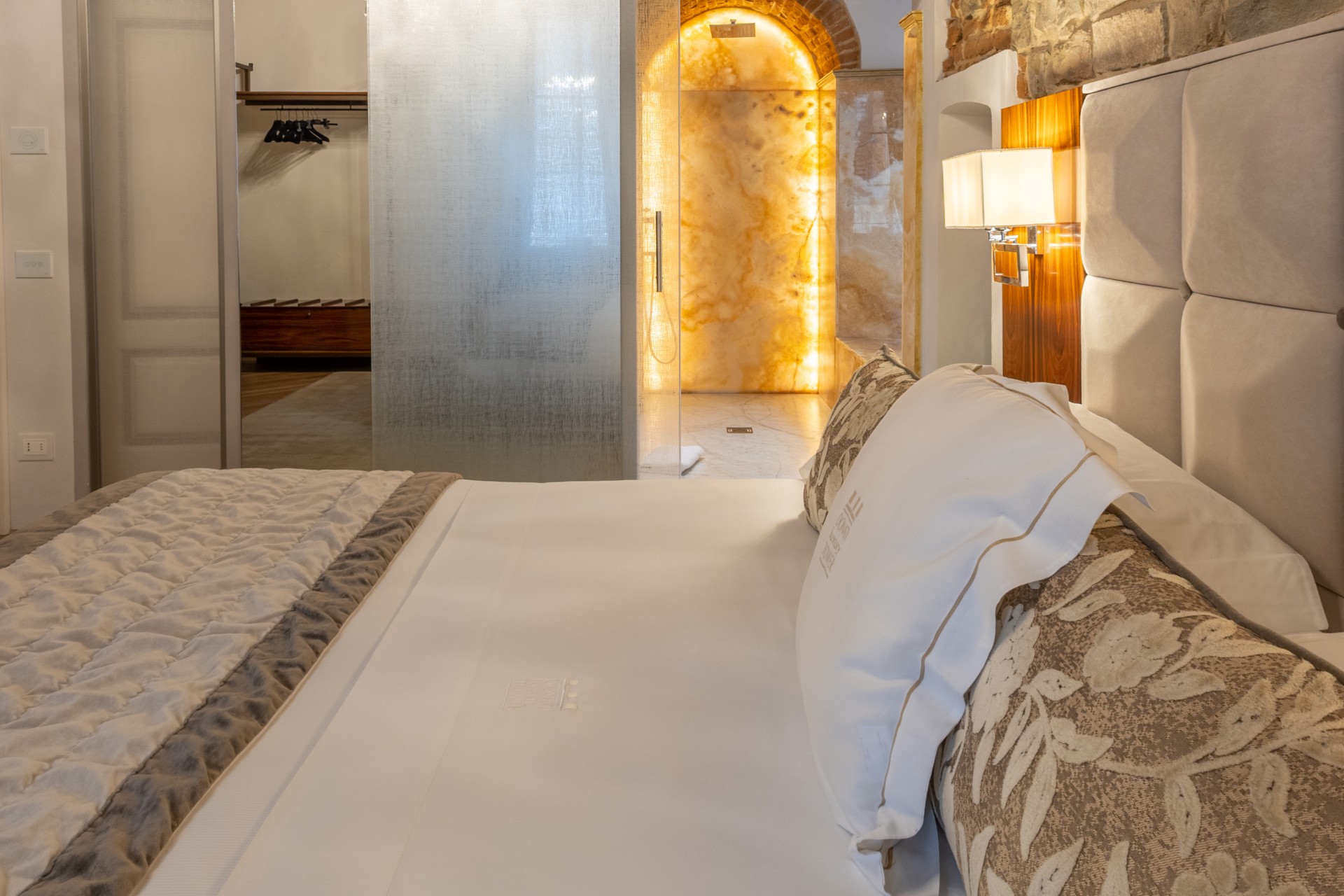 Side perspective of a hotel bedroom suite bed looking toward a glass bathroom partition with back-lit onyx wall and an open closet area.