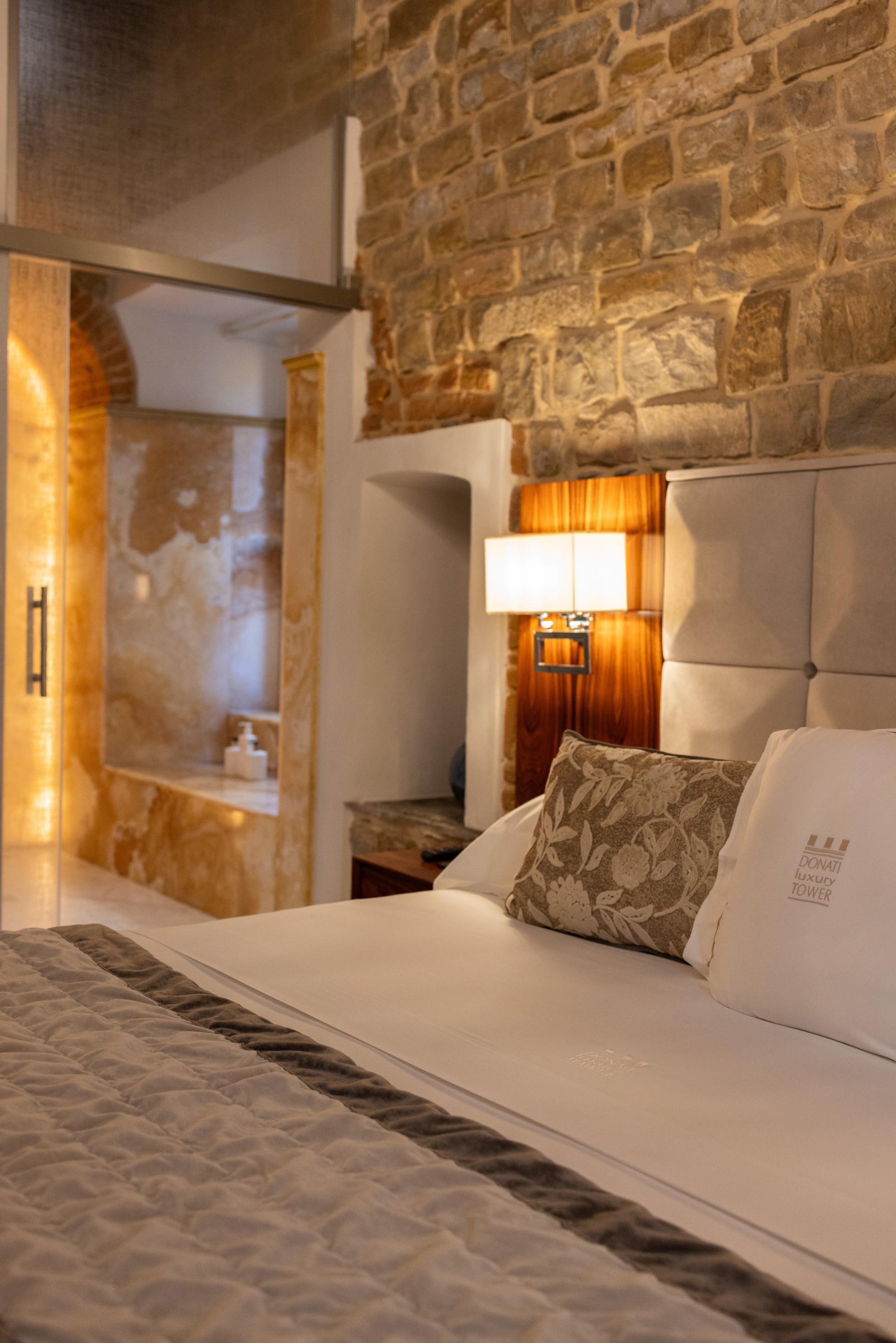 Hotel suite bed with branded pillow set against an exposed stone wall, featuring a view into an en-suite bathroom with illuminated marble finishes.