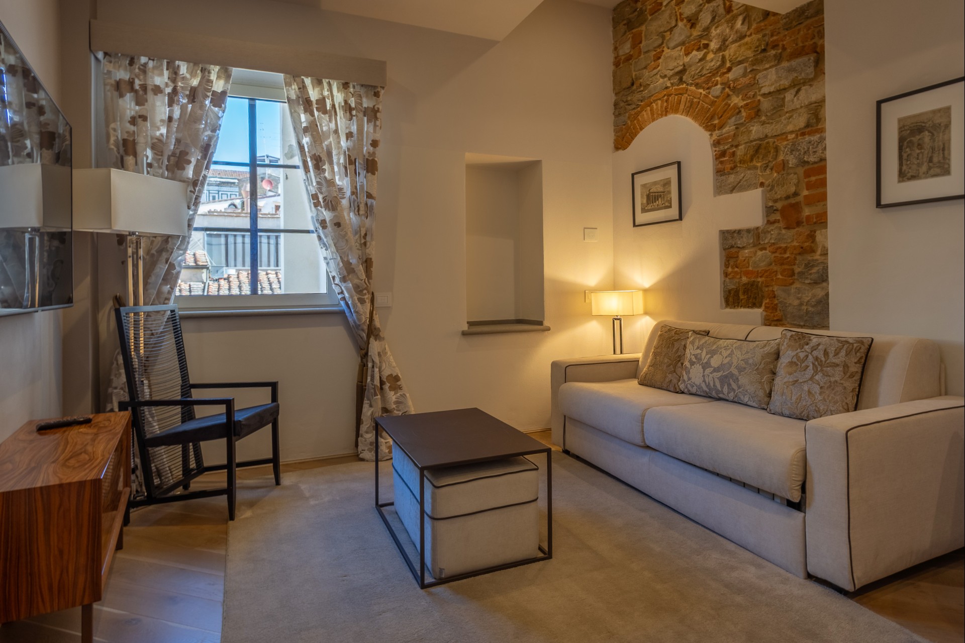 Cozy hotel suite living room with a light cream sofa, coffee table with storage ottoman, modern black accent chair, exposed brick arch, and window overlooking European rooftops.