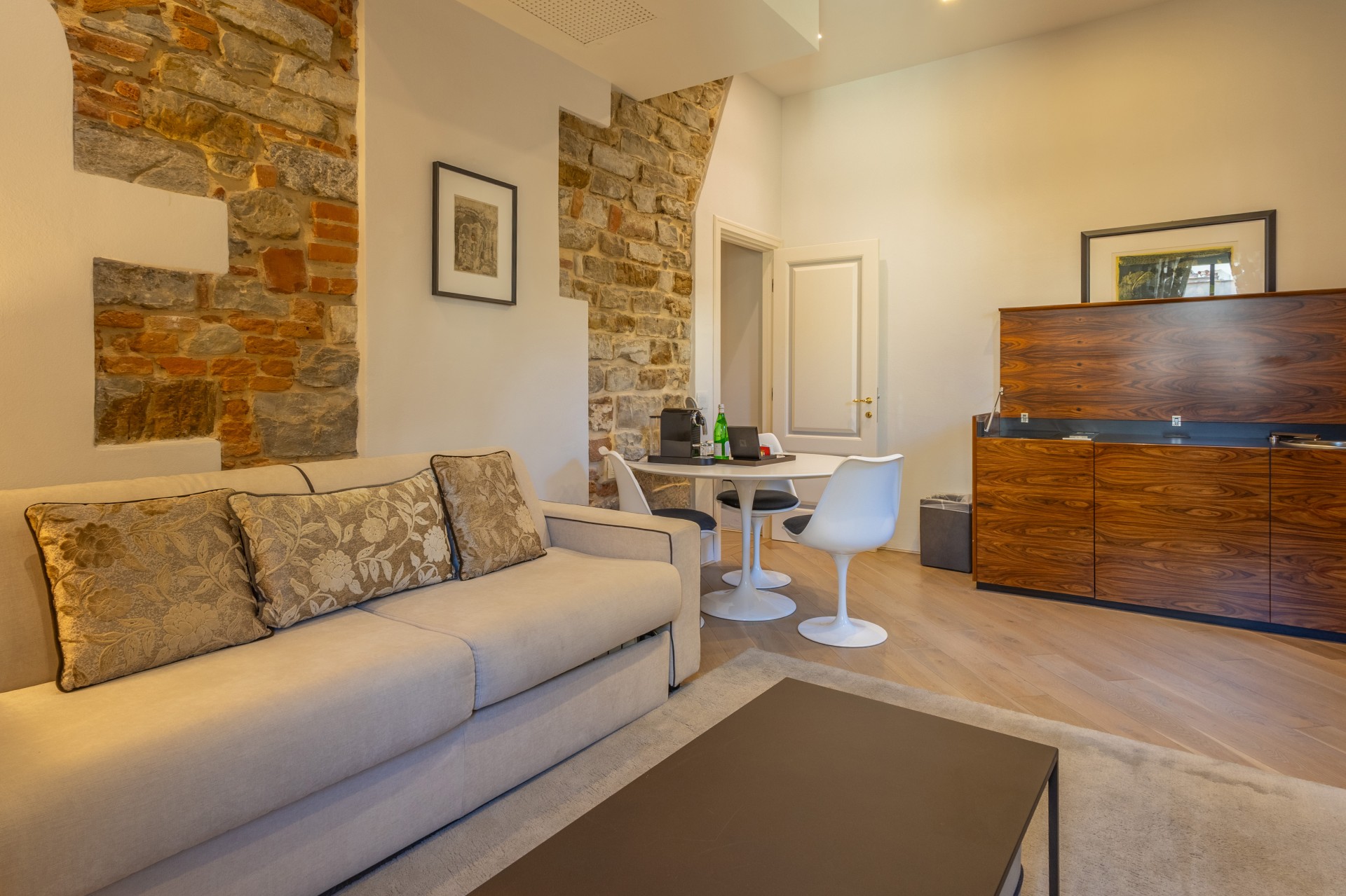 Spacious suite living area featuring a beige sofa, coffee table, white tulip dining table with modern chairs, kitchenette console, and rustic exposed stone wall accents.