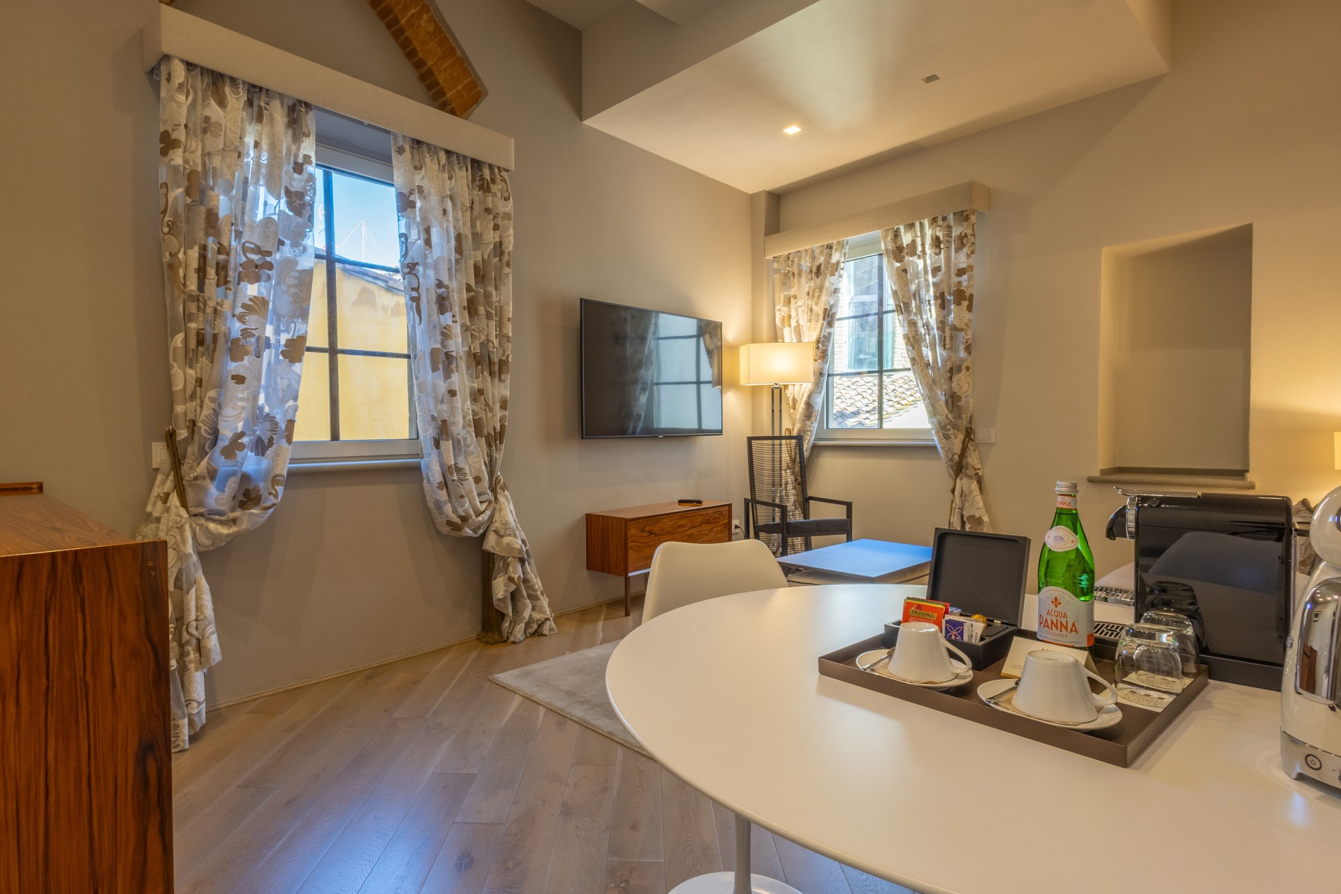 Bright hotel suite living area with a white round dining table set with tea cups and water, two large windows with patterned drapes, and a wall-mounted flat-screen TV.