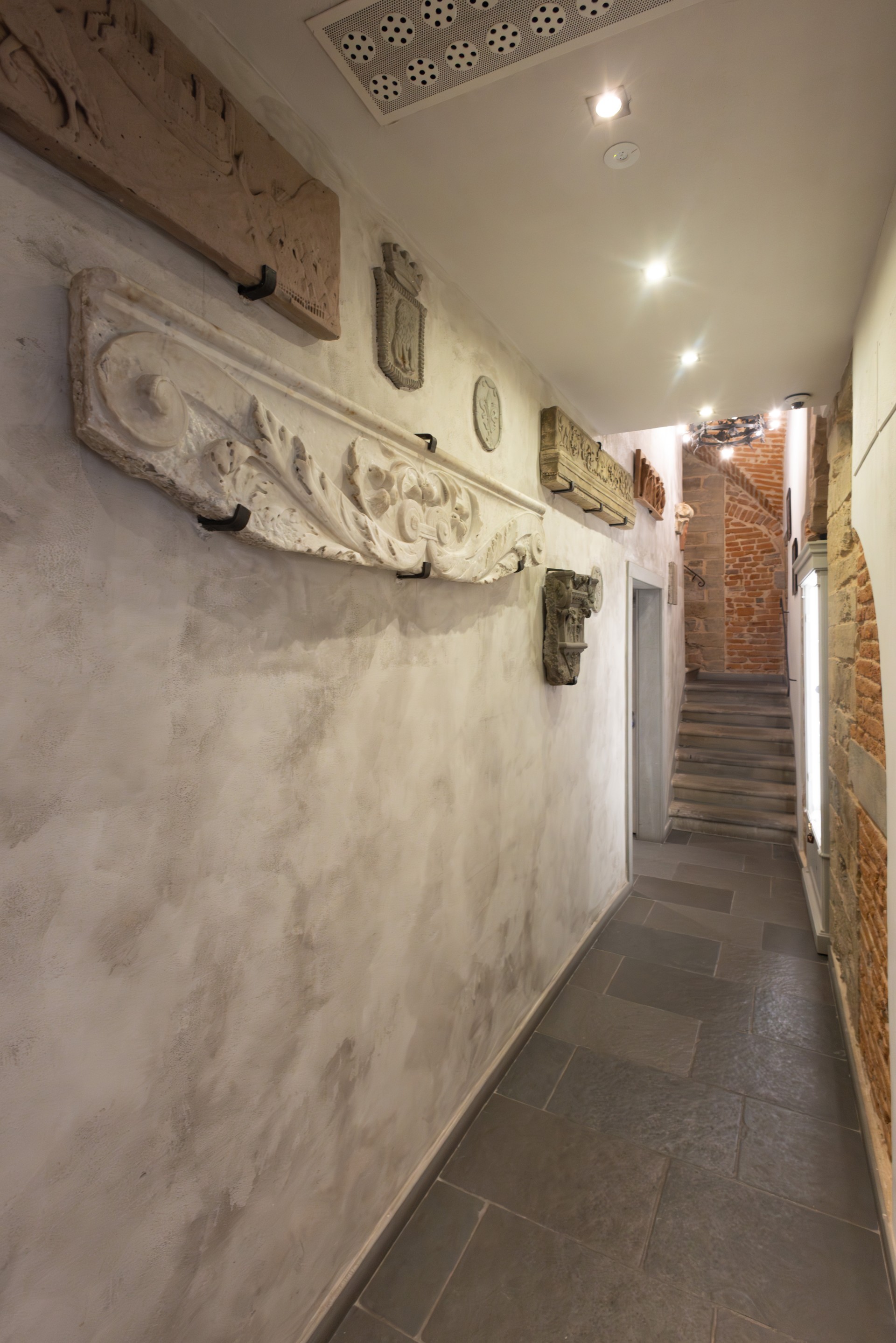 Narrow hallway in a historic Florentine building, featuring stone-tiled flooring, light plaster walls decorated with carved stone artifacts and coats of arms, leading toward a brick staircase.