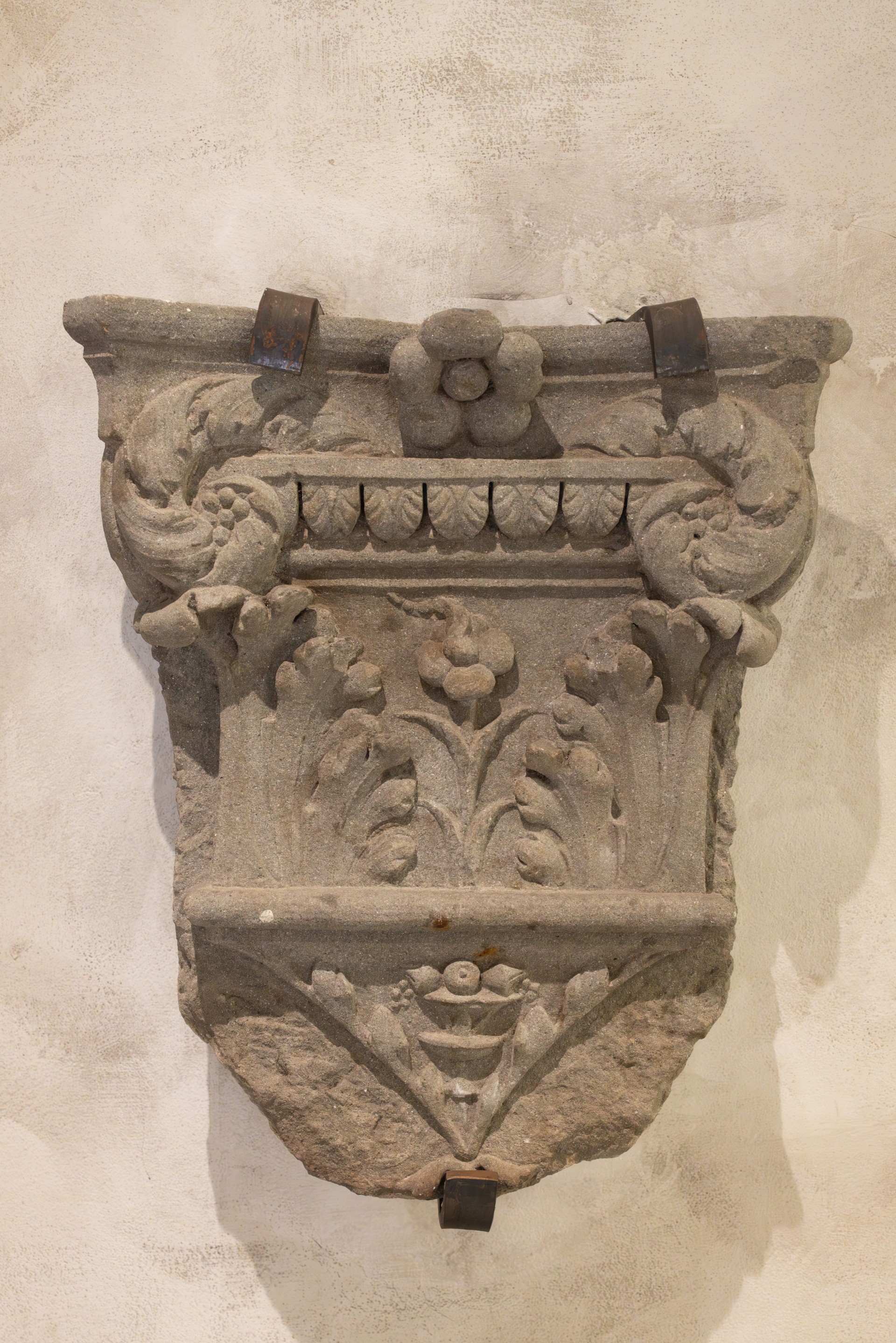 Carved grey stone corbel with decorative acanthus leaves and floral reliefs mounted on a neutral wall using metal brackets.