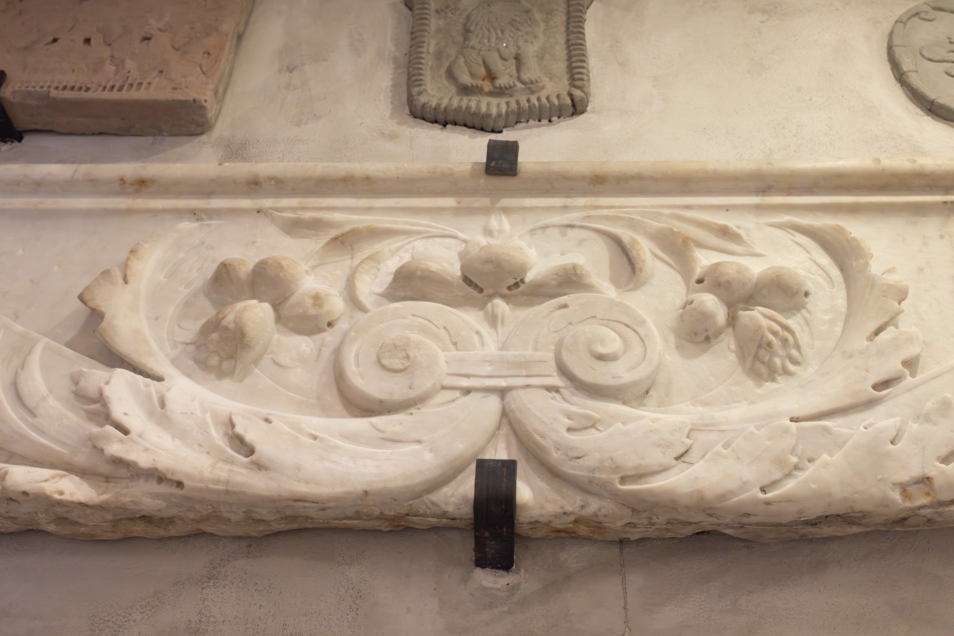 Close-up detail of an ornate white marble frieze featuring intricate carved acanthus leaves, floral motifs, and central scrolls mounted on a light wall.