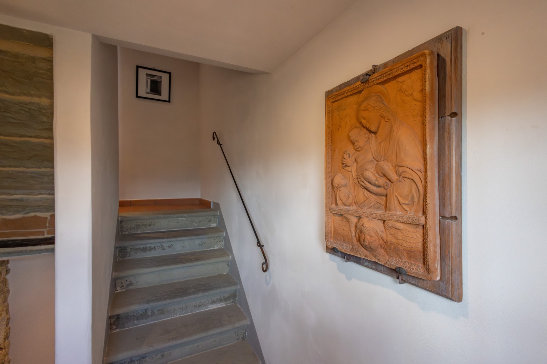 Narrow indoor staircase with grey stone steps and a simple wrought-iron handrail, featuring a terracotta relief plaque of the Madonna and Child on the adjacent white wall.
