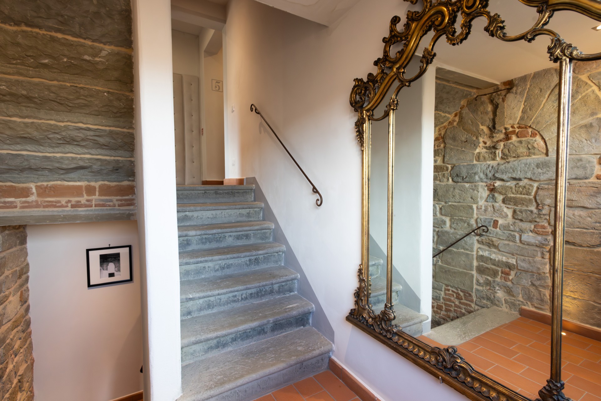 Interior staircase area with grey stone steps, terracotta floor tiles, exposed stone brickwork, and a large ornate gilt-framed mirror reflecting the landing.
