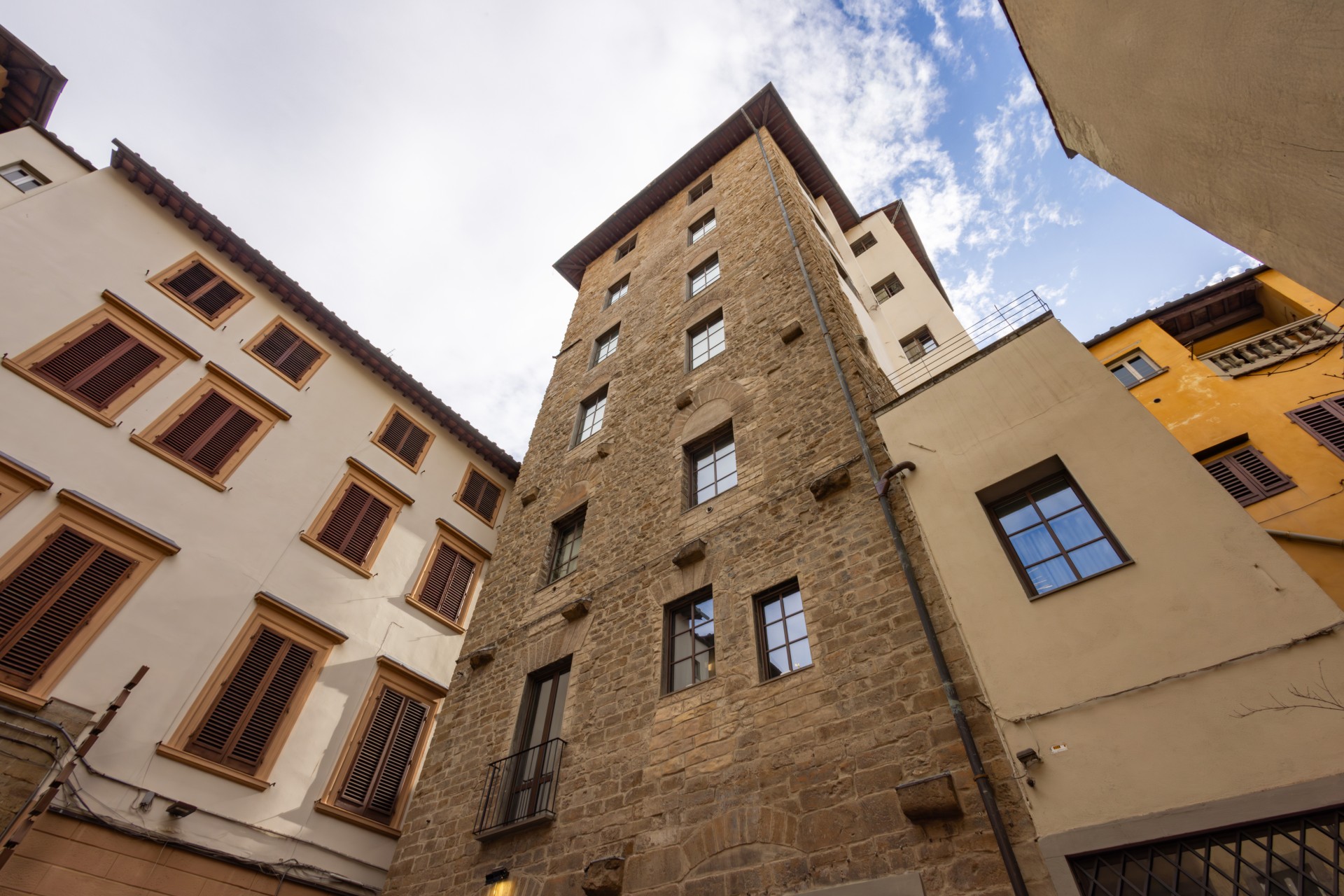 Worm's-eye view looking up at a historic stone tower in Florence, framed by surrounding traditional buildings under a bright sky with light clouds.