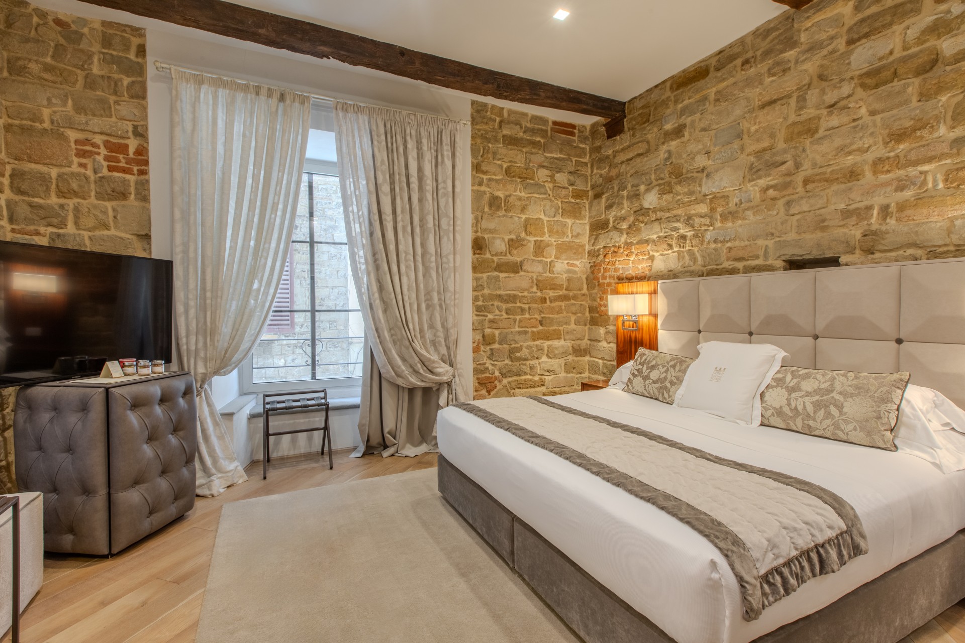 Hotel bedroom featuring exposed stone walls, exposed wooden ceiling beams, a large king bed with a tufted headboard, a window with sheer curtains, and a television.